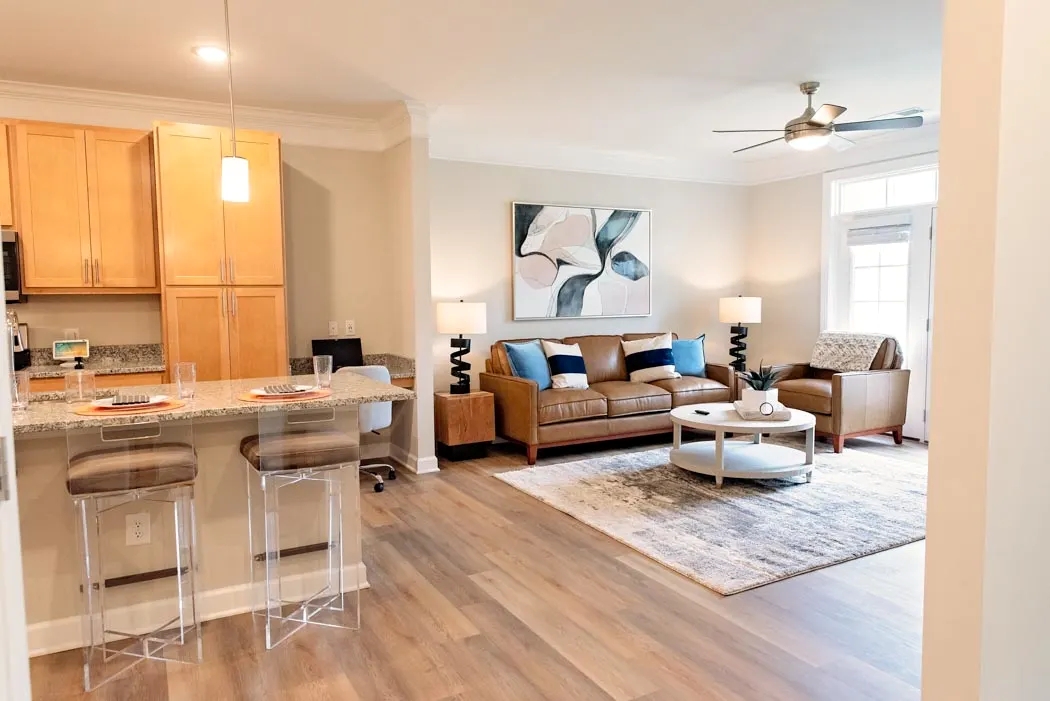 An open-concept apartment living area showing a leather sofa, breakfast bar with acrylic stools, and wood flooring.