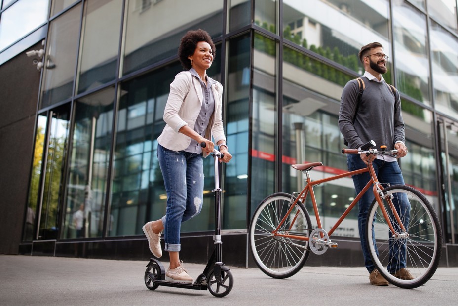 Two people outside a modern glass building, one riding a scooter and the other walking with a bicycle.
