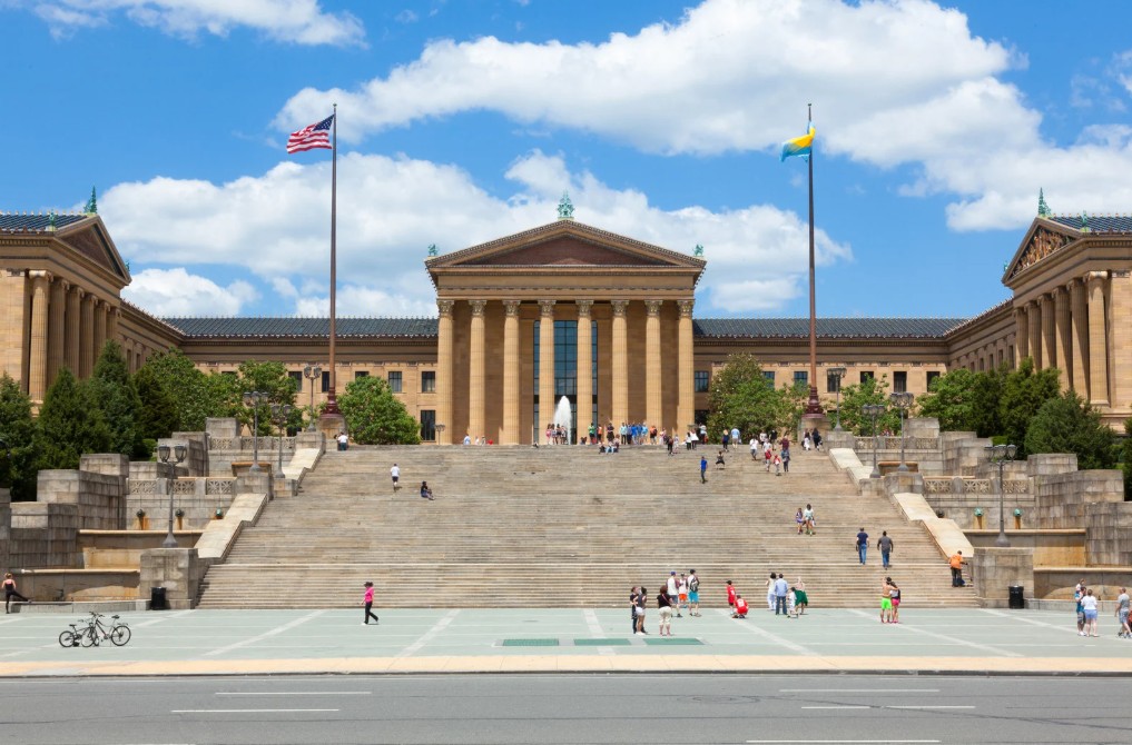 Daytime view of the Philadelphia Museum of Art with its iconic neoclassical facade, wide steps, and partly cloudy sky in the background.          Ask ChatGPT