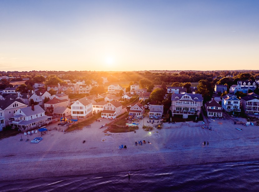 An aerial sunset view of beachfront homes along the coastline in Fairfield, Connecticut, with golden light reflecting off the water and sand.