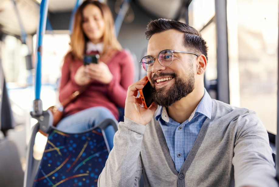A smiling, bearded man wearing glasses is talking on a smartphone while riding a bus in Fairmount, with a woman looking at her phone seated in the blurry background.