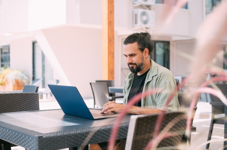 A bearded man with a top knot working on a blue laptop at an outdoor table in a sunny urban setting in Fairmount, Philadelphia.