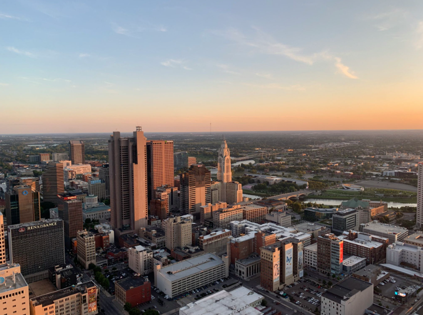 Aerial view of the Downtown Columbus skyline at sunset, featuring the city's high-rise buildings, the Scioto River, and surrounding Central Ohio neighborhoods.