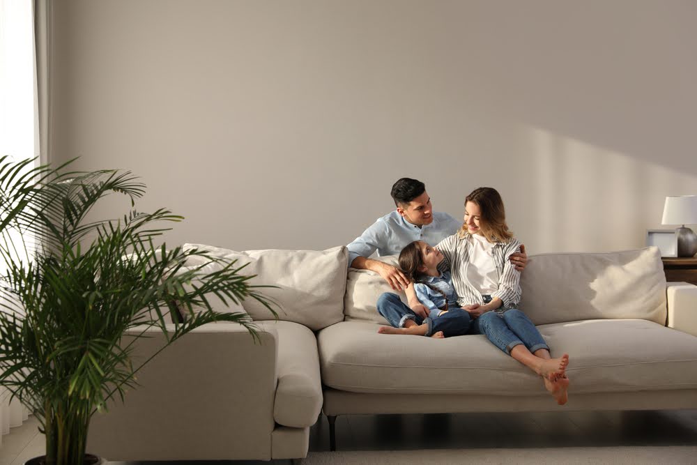 A smiling family relaxes on a beige sofa in a bright, cozy living room at Wesley Hampstead at Douglasville in Lithia Springs, GA.