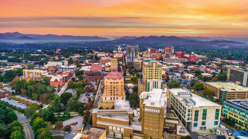 Aerial view of Asheville, NC at twilight, with city buildings and mountains under a warm, colorful sky.