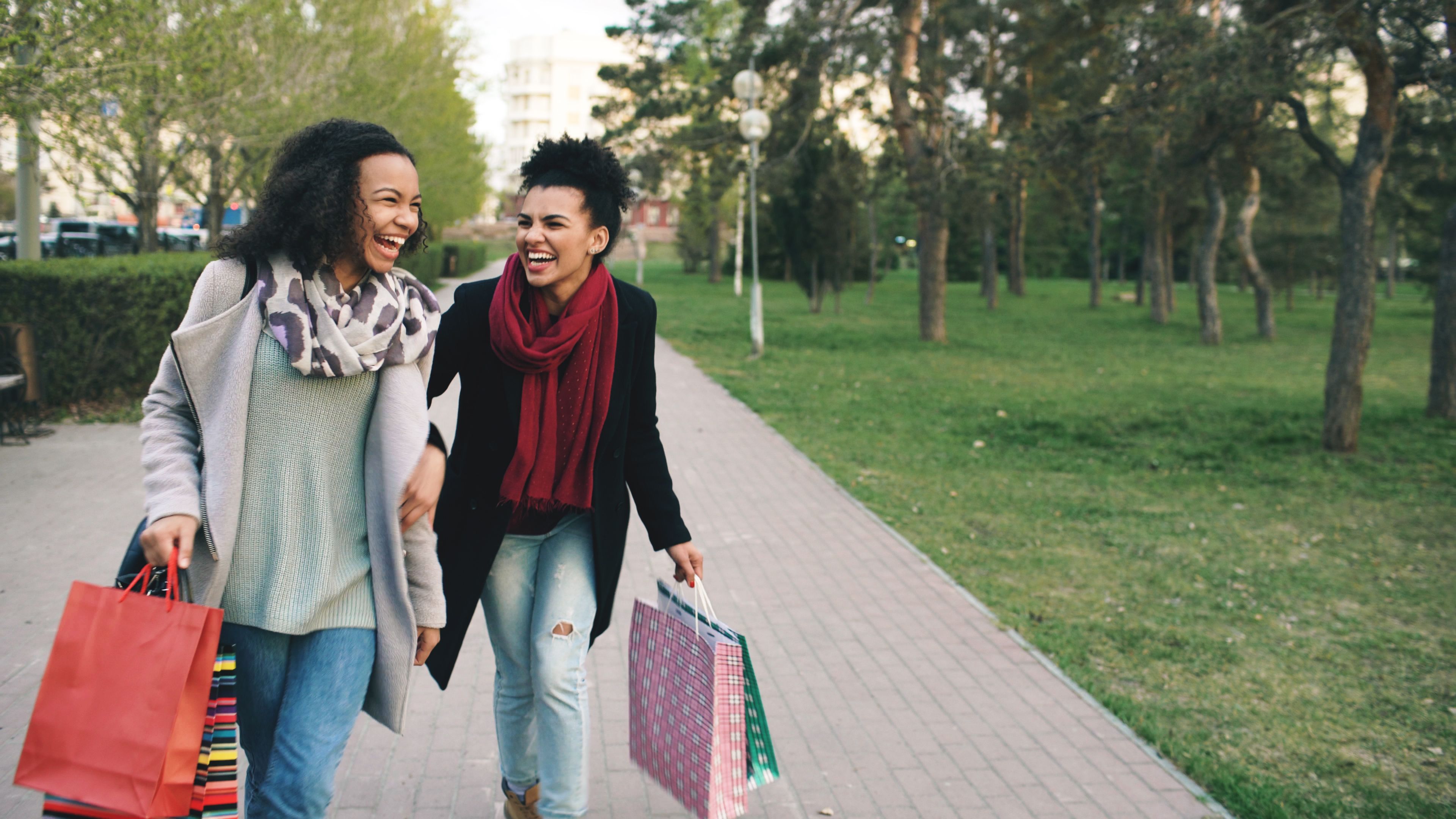 Two laughing women wearing coats and scarves carrying colorful shopping bags walking on a paved path next to trees.