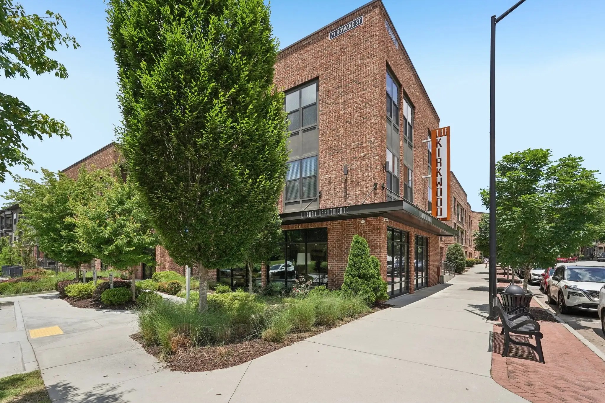 A brick apartment building displaying a vertical The Kirkwood sign standing near a concrete sidewalk and trees.
