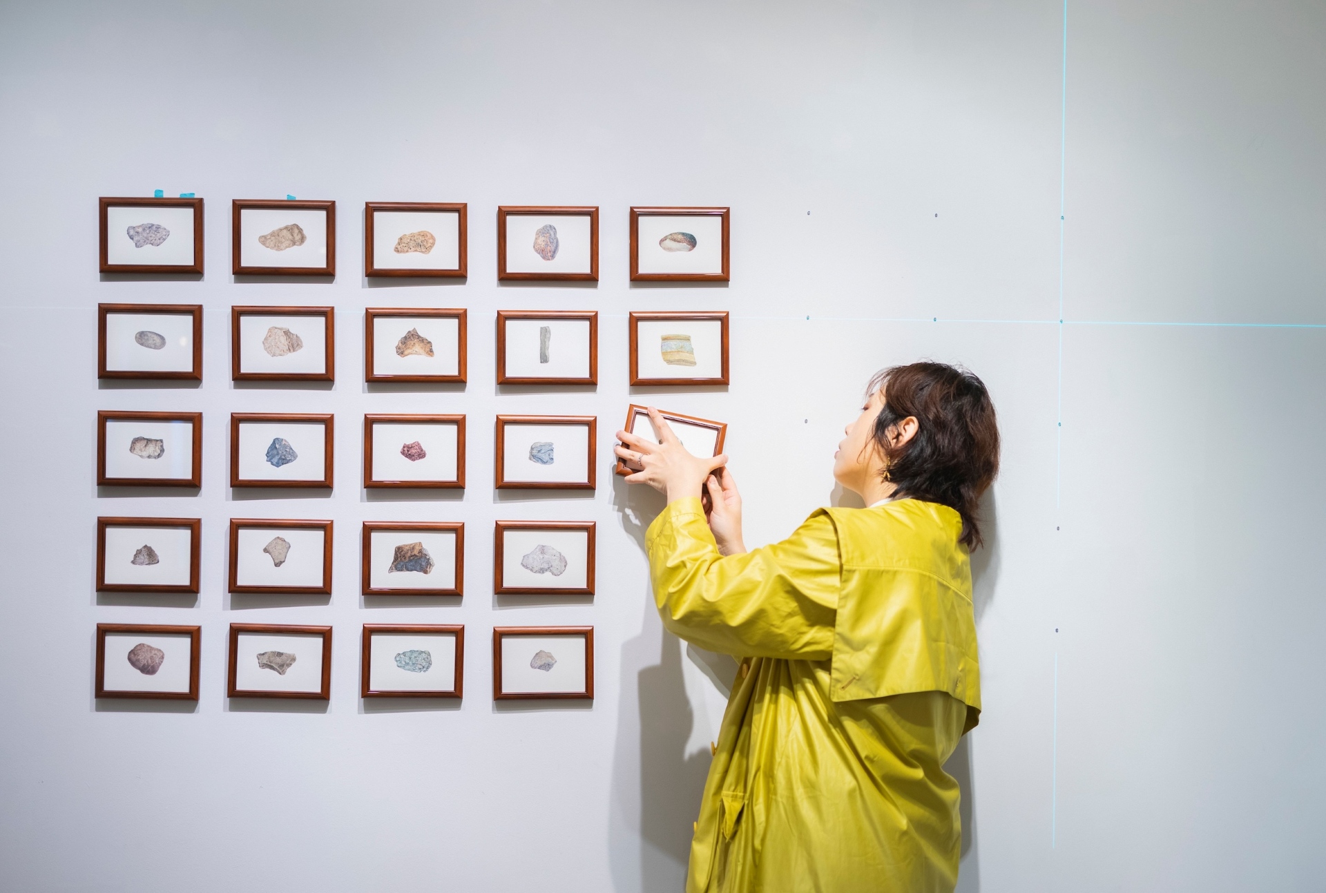A woman wearing a yellow coat standing near a white wall hanging a small framed picture next to a grid of frames