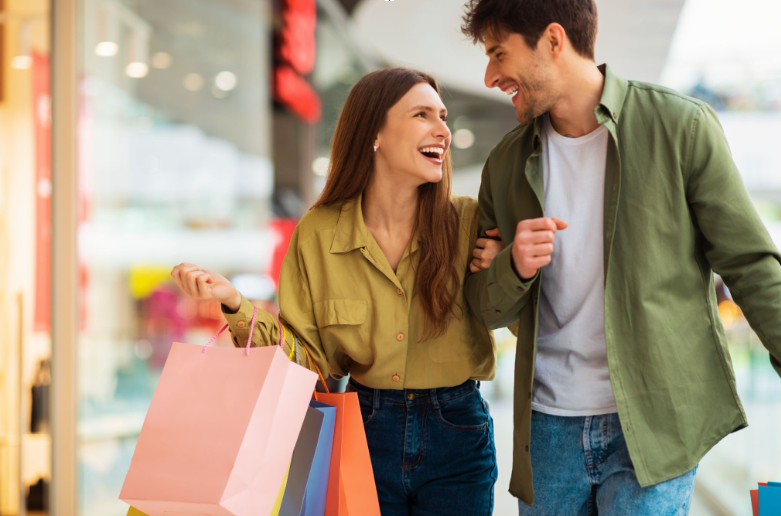 A happy young couple walking arm-in-arm, laughing, and carrying colorful shopping bags inside a mall or outlet center in Foley, Alabama.