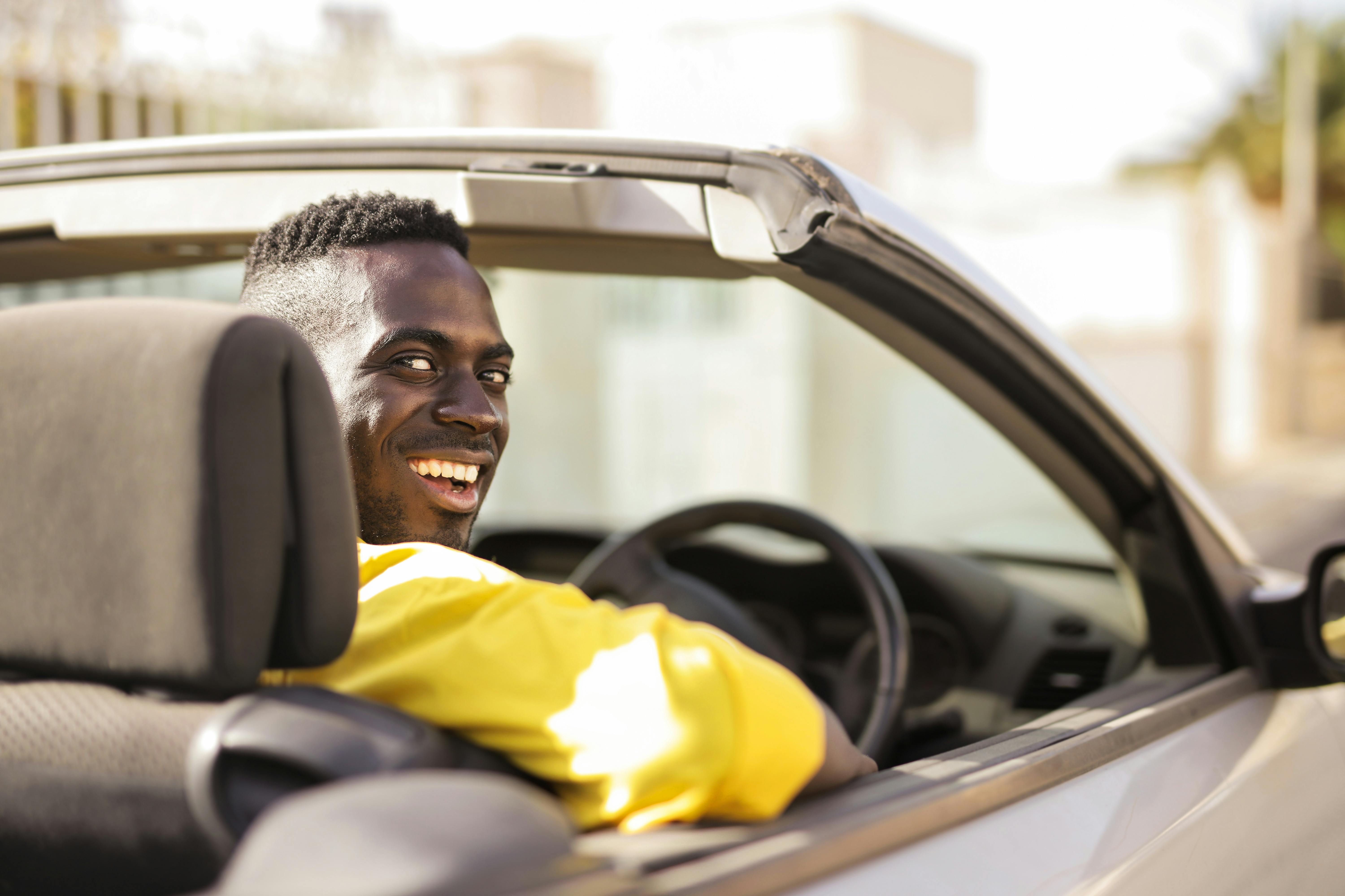 A smiling man wearing a yellow shirt sitting inside a white convertible car looking back along a sunny street.
