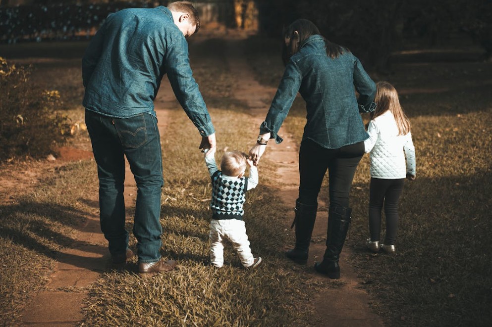 Family walking hand in hand along a park path, with parents guiding a toddler and an older child.