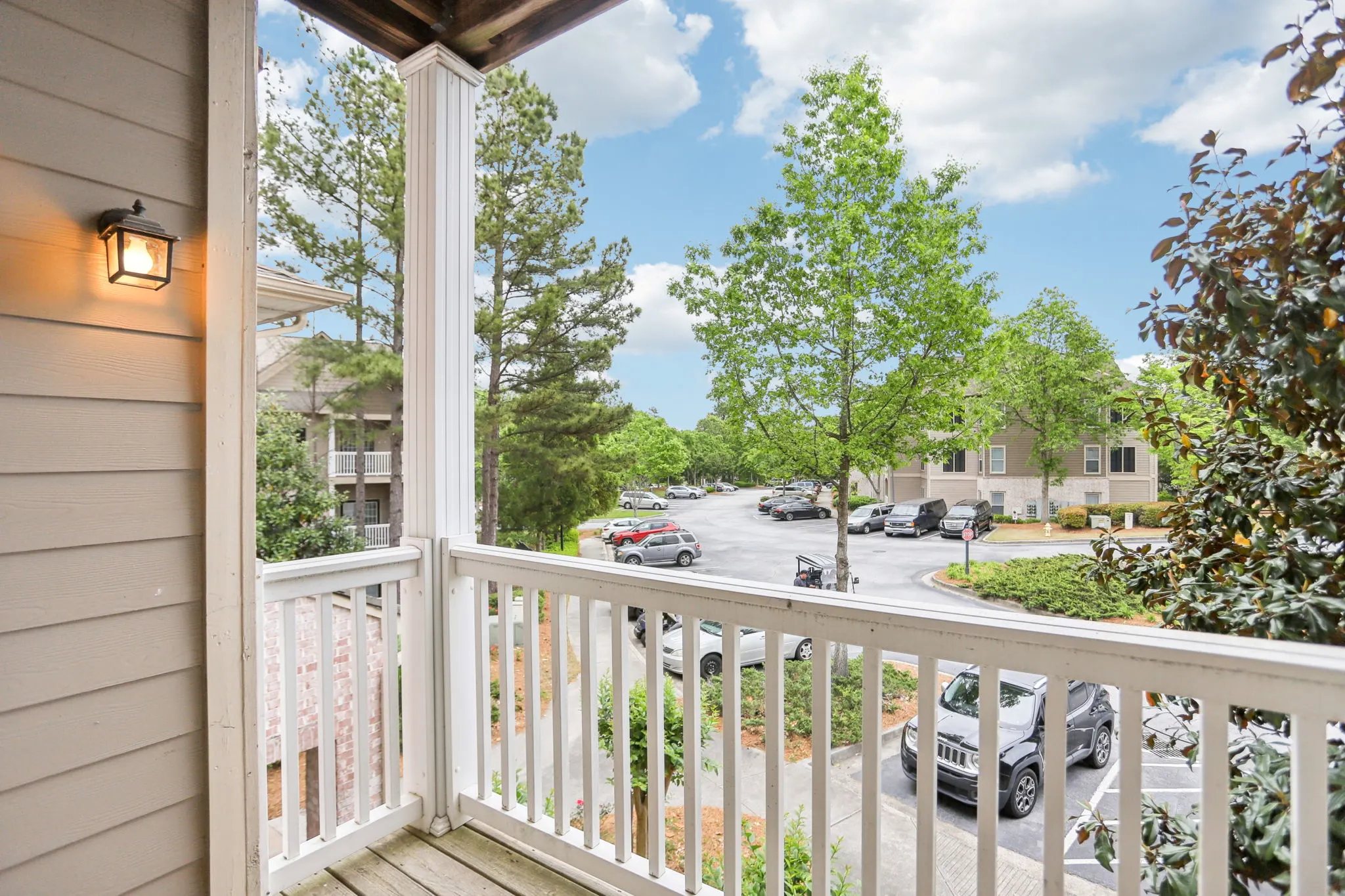 A private apartment balcony featuring white railings and an outdoor light overlooking a parking lot and trees.