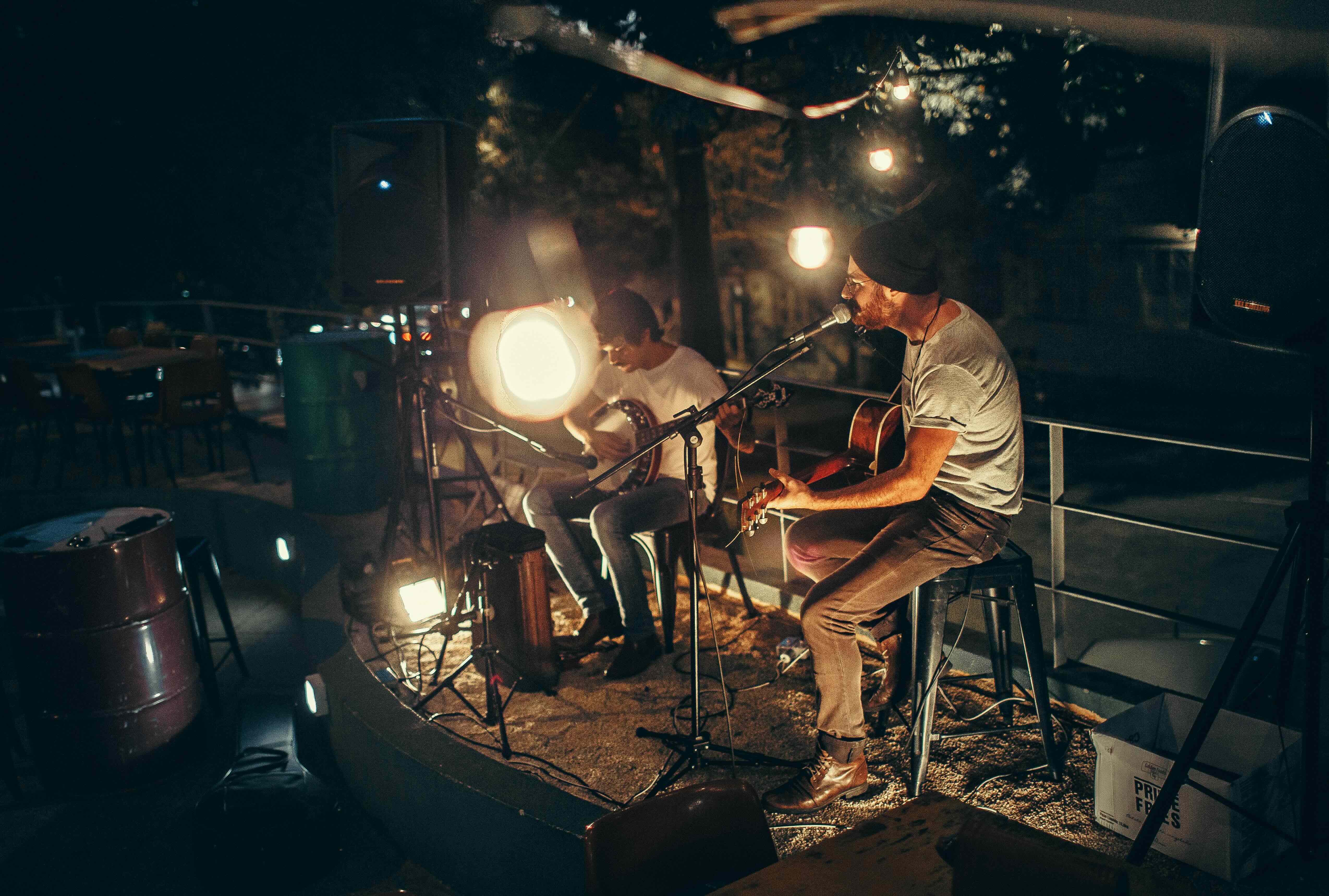 Two men wearing casual clothing sitting on stools playing stringed instruments and singing on a lit outdoor stage.