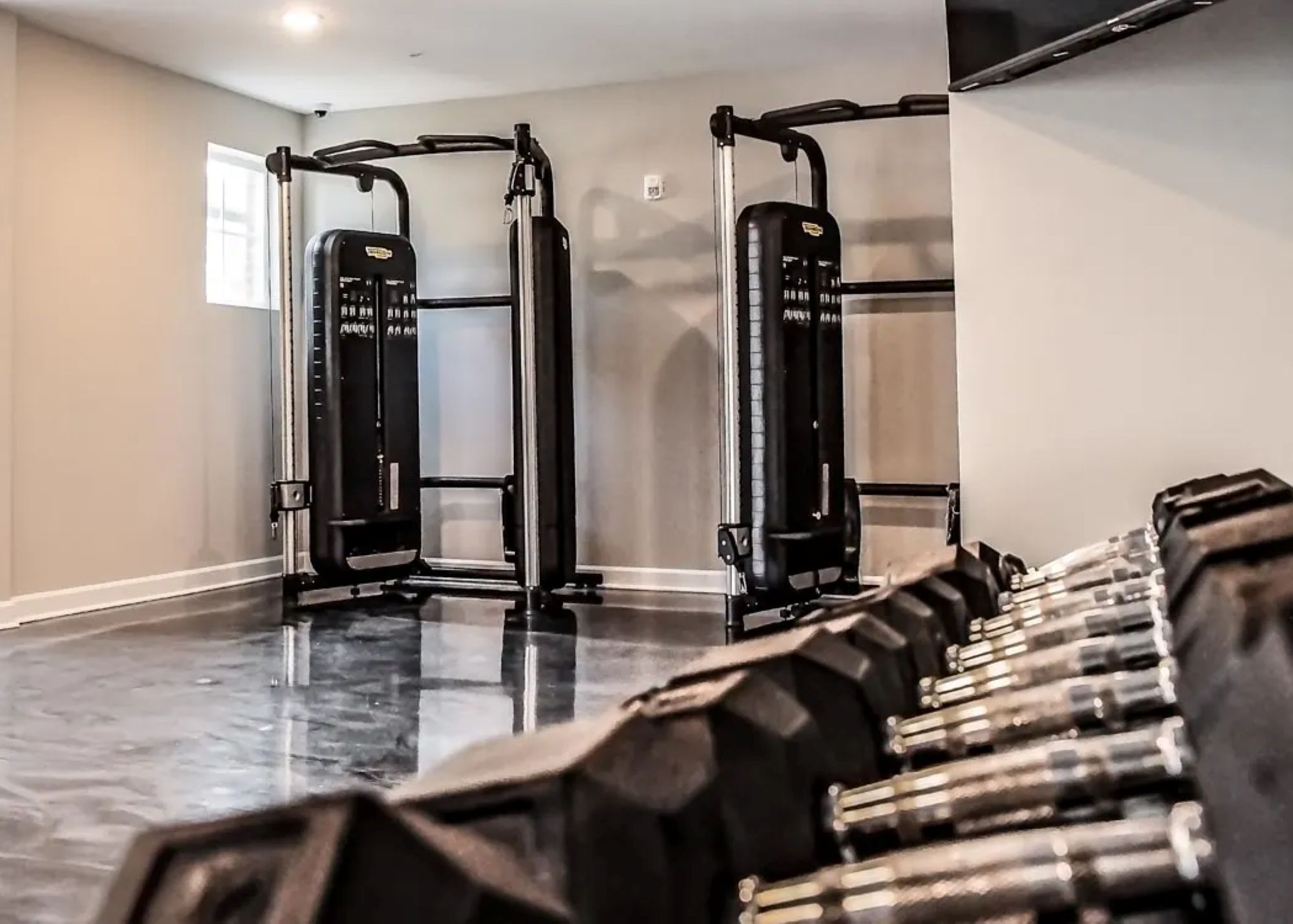 An apartment fitness center featuring cable weight machines and a row of dumbbells on a glossy dark floor.