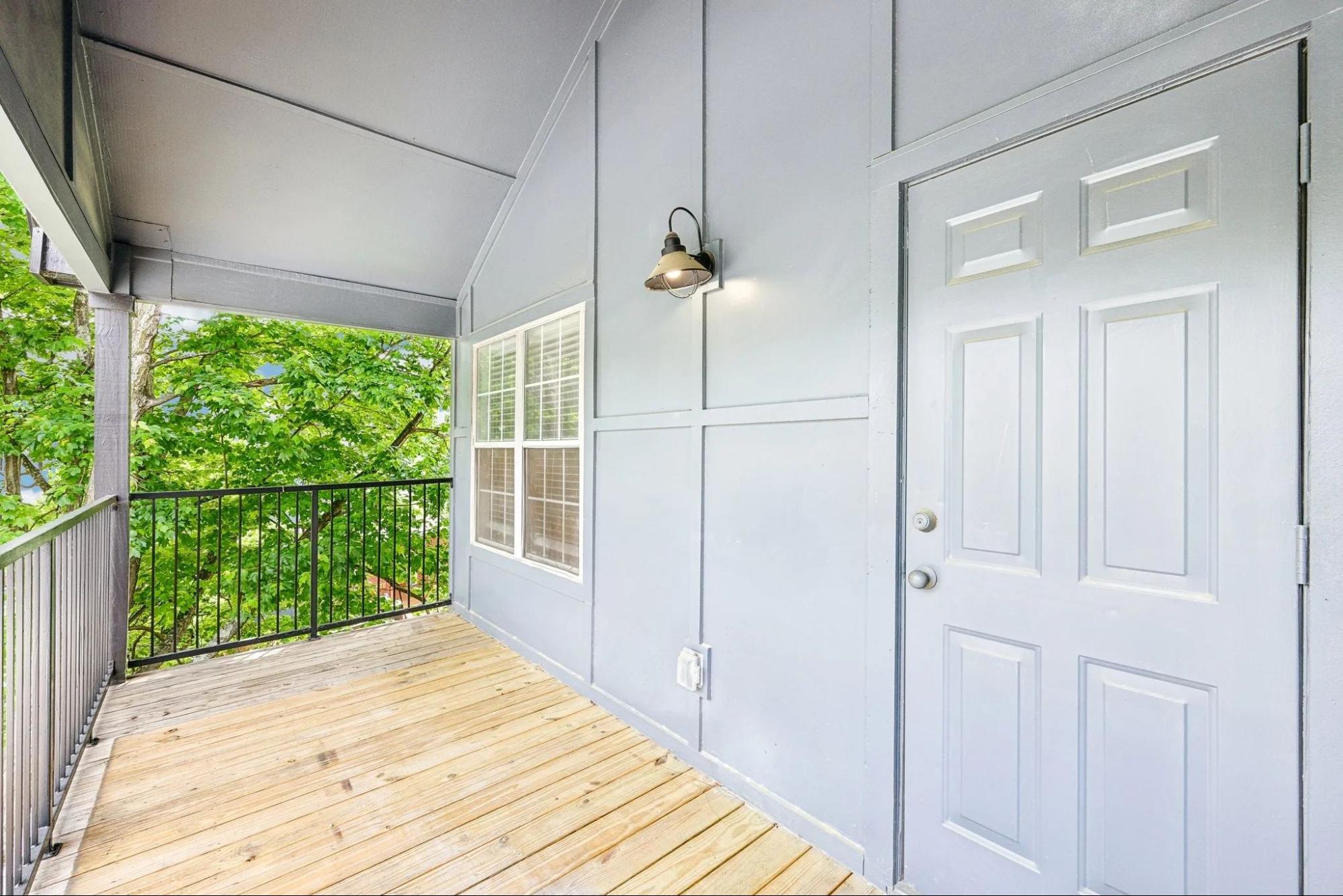 A covered apartment balcony featuring wooden deck flooring, a grey door, and views of lush green trees.