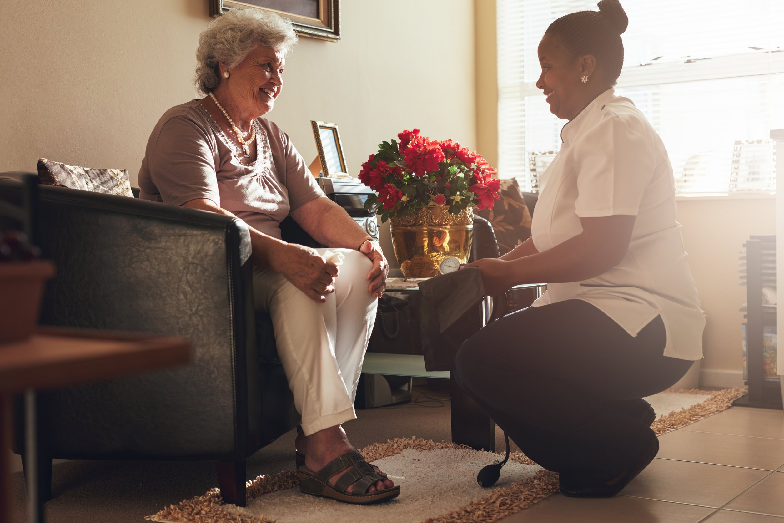 Senior woman sitting on a chair at home with female caregiver holding blood pressure gauge