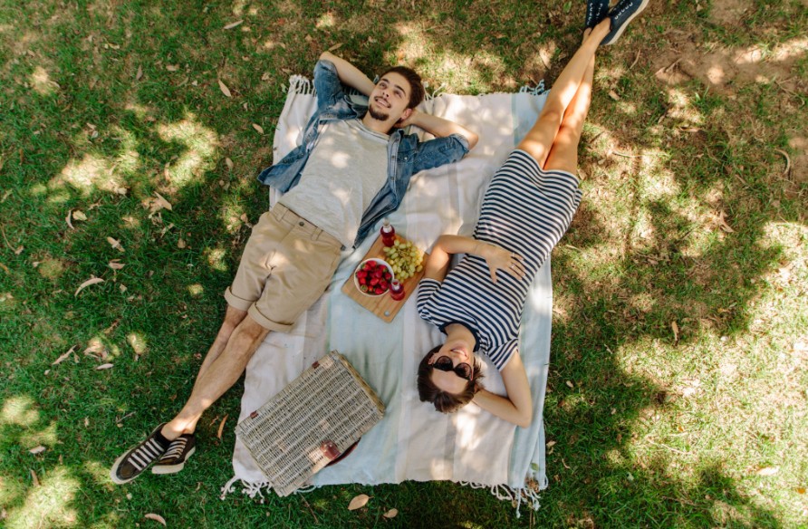 Couple lying on picnic blanket in shaded grass with fruit and wicker basket
