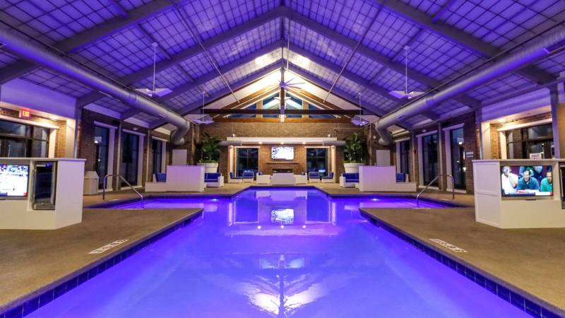 Luxury indoor pool area at Kensington at Riggins Run Apartments featuring lounge seating, mounted TVs, and ambient purple lighting.