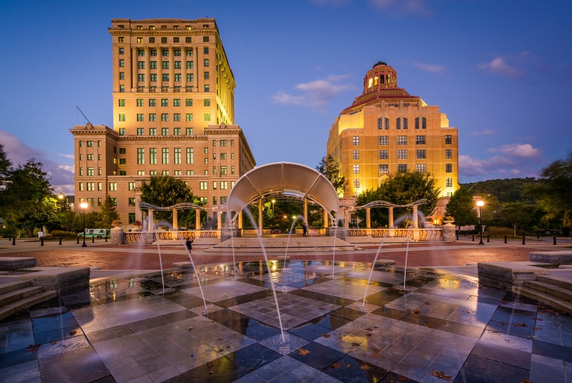 Pack Square Park fountain in Asheville, NC at dusk, with the illuminated City Hall and County Courthouse visible in the background.
