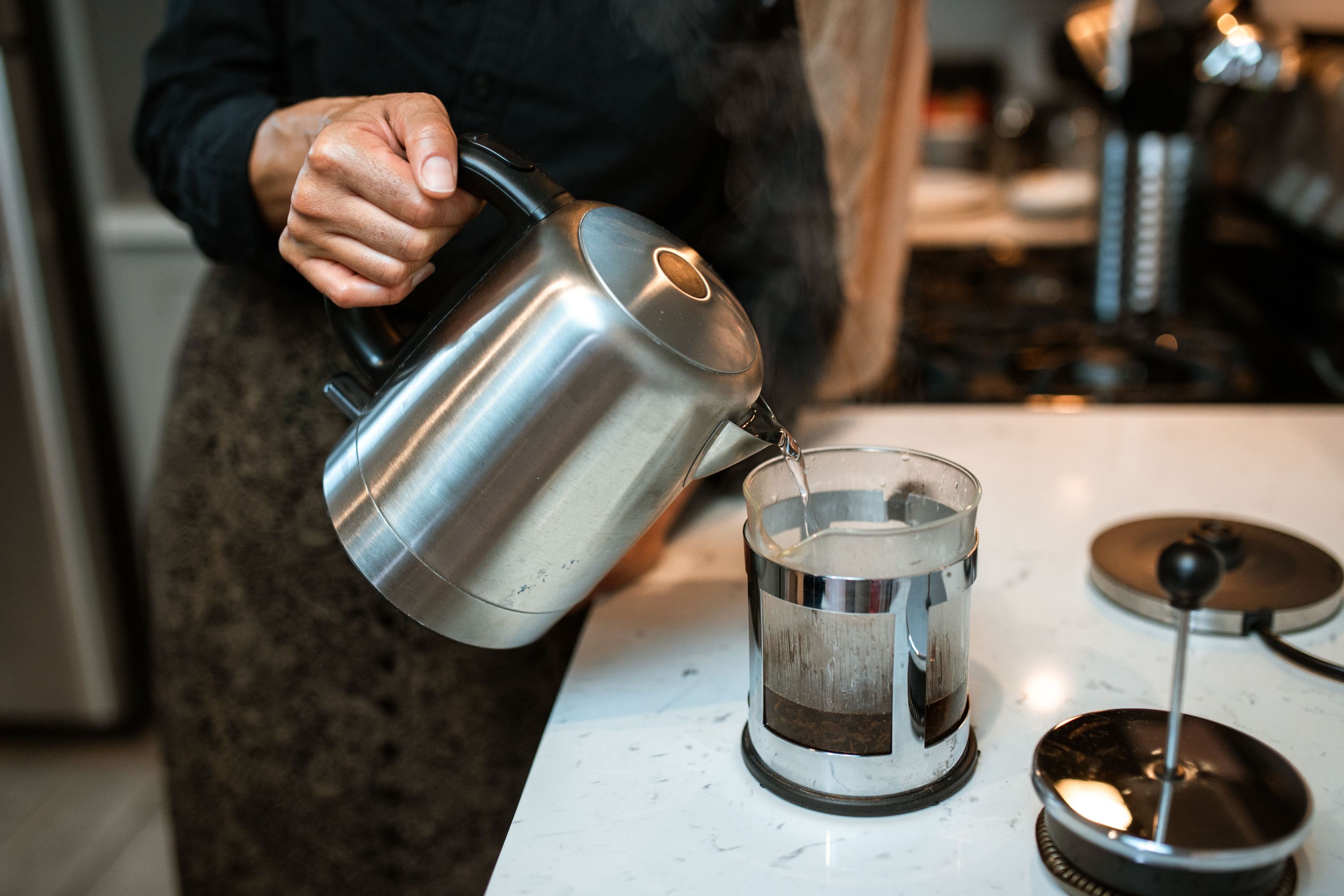 A person pouring hot steaming water from an electric stainless kettle into a French press on a marble counter.