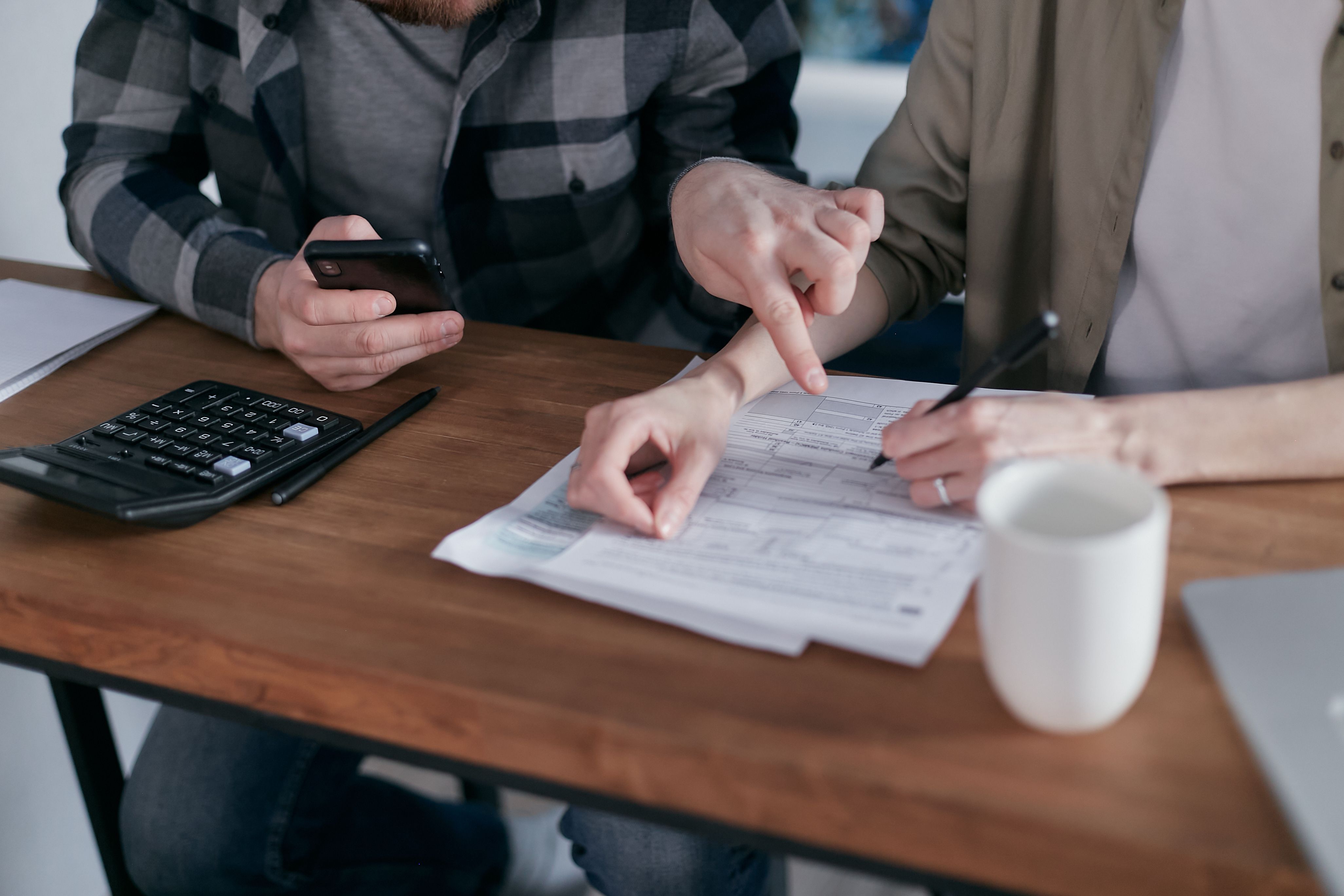 A couple reviewing financial documents while filling out paperwork at a wooden desk with a calculator and phone.