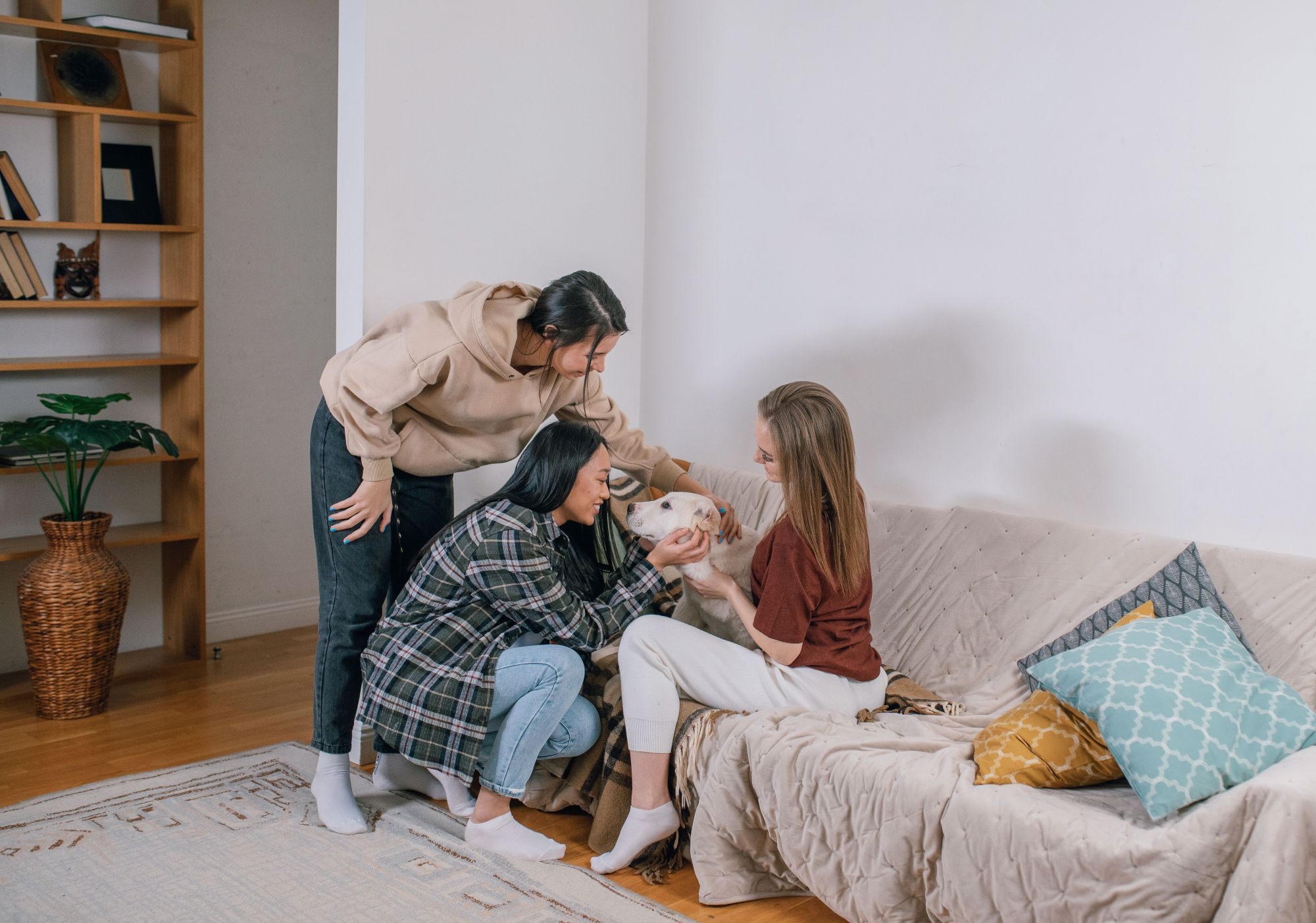 Three women smiling and petting a large white dog resting on a beige couch inside a room with a wooden bookshelf.