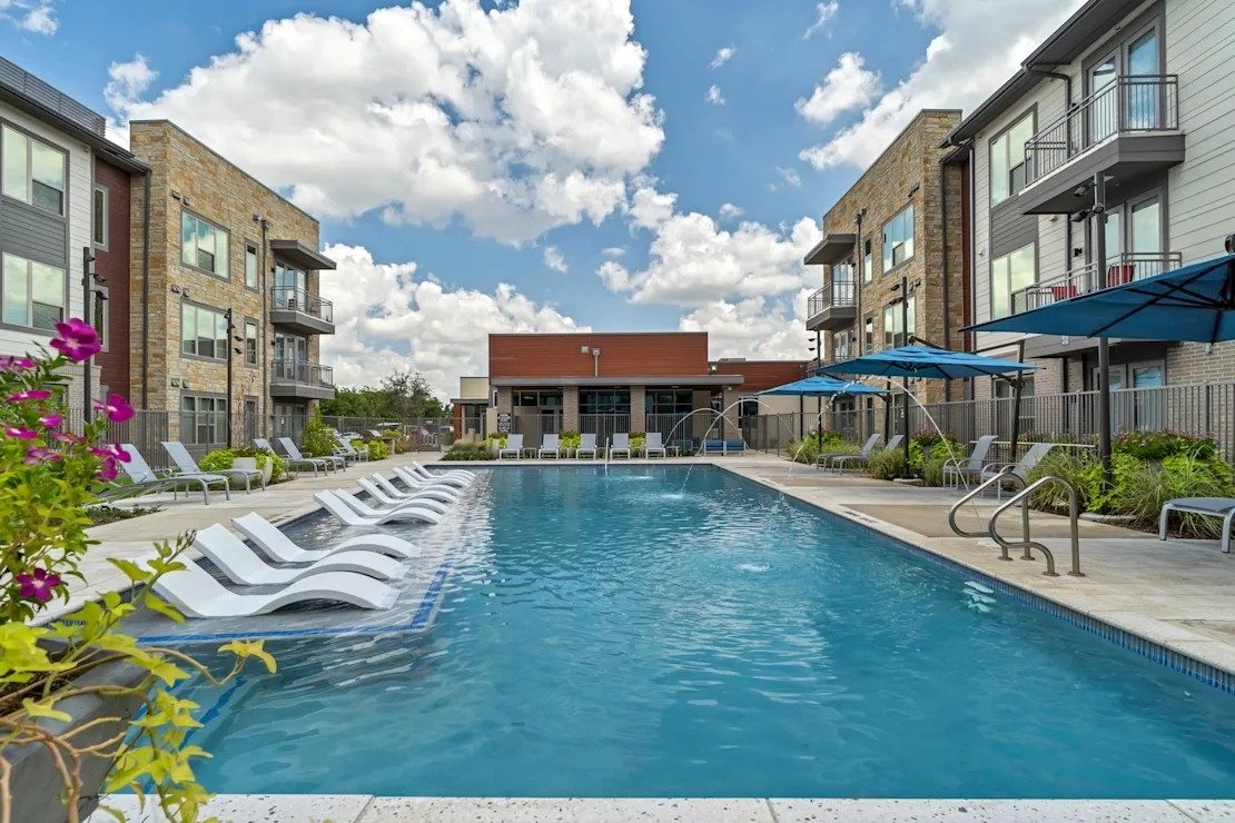 A rectangular swimming pool featuring white lounge chairs and blue umbrellas flanked by two modern apartment buildings