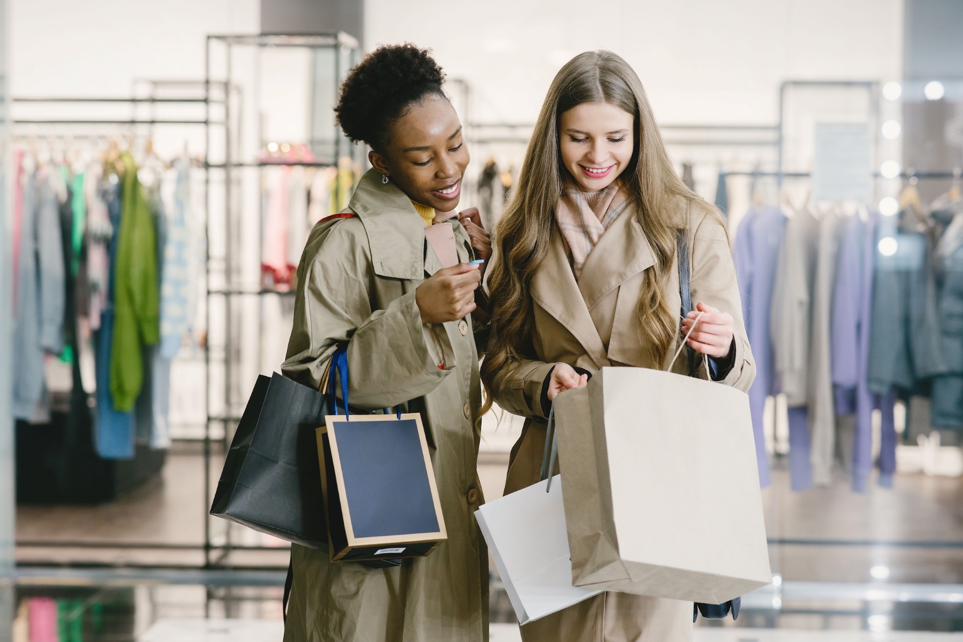 Two smiling women wearing tan coats standing inside a retail store looking into a large paper shopping bag