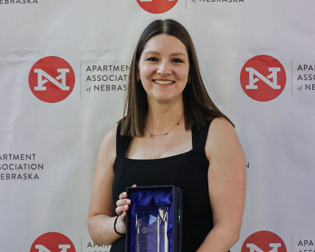 Corryn Wilson holding her award in front of a backdrop with the Apartment Association of Nebraska logos on it