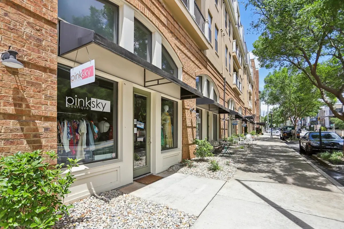 A brick storefront displaying a pinksky boutique sign resting near a concrete sidewalk lined with green trees.