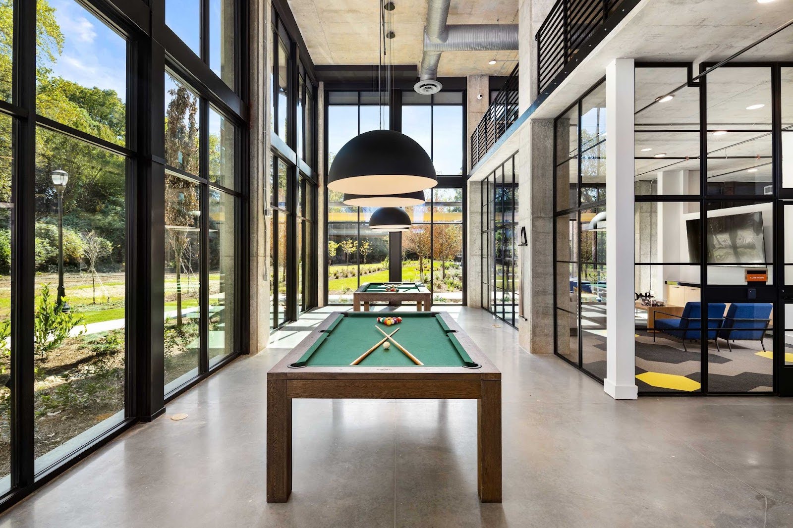 A pool table in a modern, sunlit interior space, featuring large windows and an industrial-style concrete ceiling at The Argos Apartments.