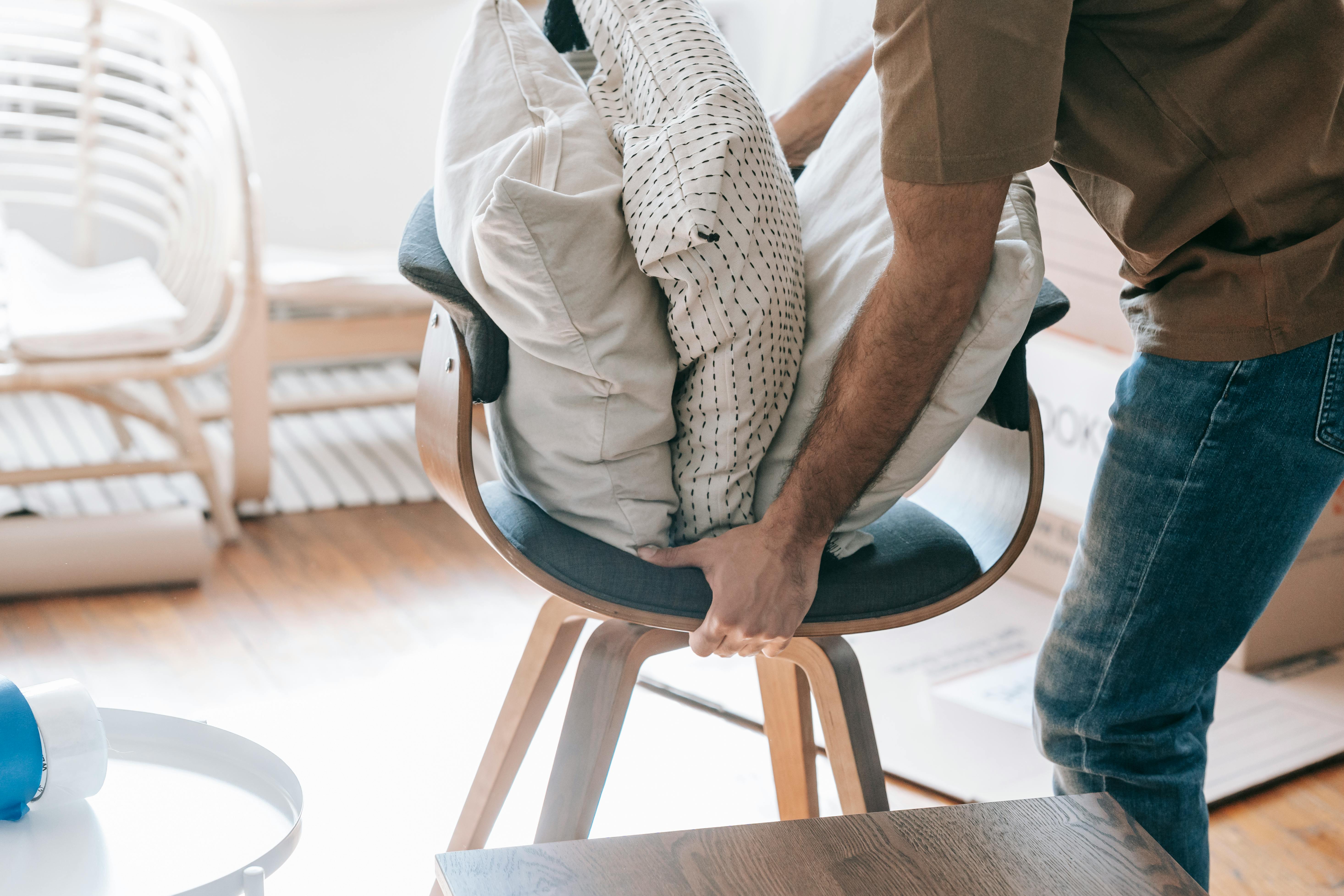 A person wearing blue jeans and a brown shirt placing light pillows onto a wooden chair near a cardboard box.