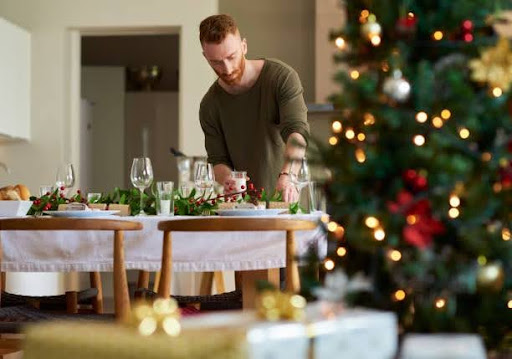 A man setting a festive dining table with wine glasses and a holiday garland, positioned next to a decorated holiday tree in Wesley St. Claire Apartments.