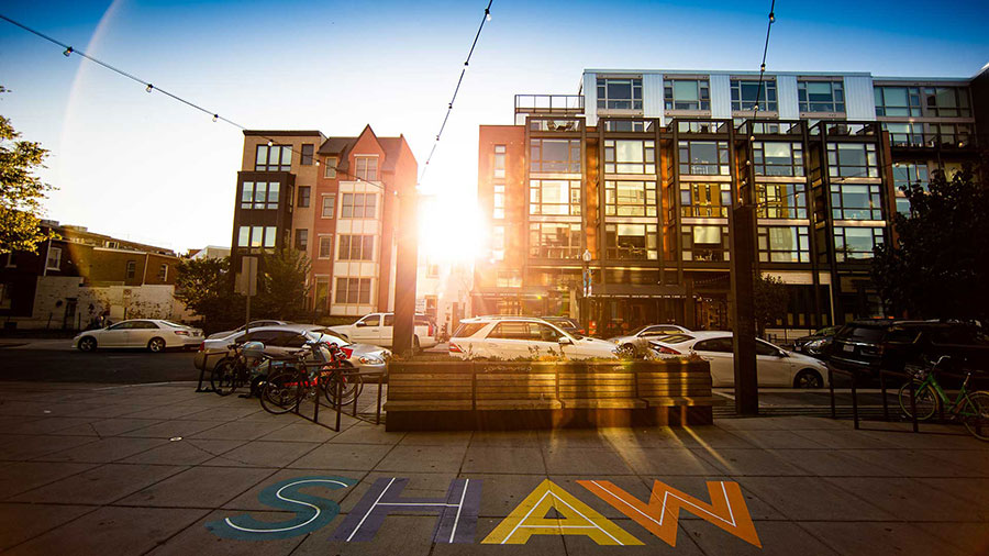 Street-level view of the Shaw neighborhood in Washington, D.C., with colorful painted letters spelling SHAW on the sidewalk, bike racks, and mixed-use residential buildings at sunset