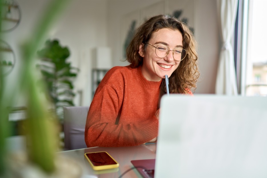 A smiling remote worker with curly hair and glasses sits at a desk in an Asheville home office, looking at a laptop and holding a pen.