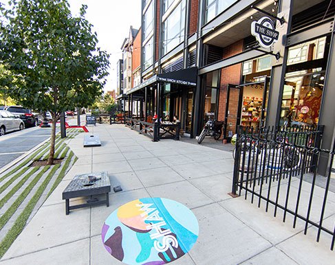 Tree-lined sidewalk in Shaw with brick storefronts, outdoor seating, cornhole boards, and a painted Shaw neighborhood graphic on the pavement