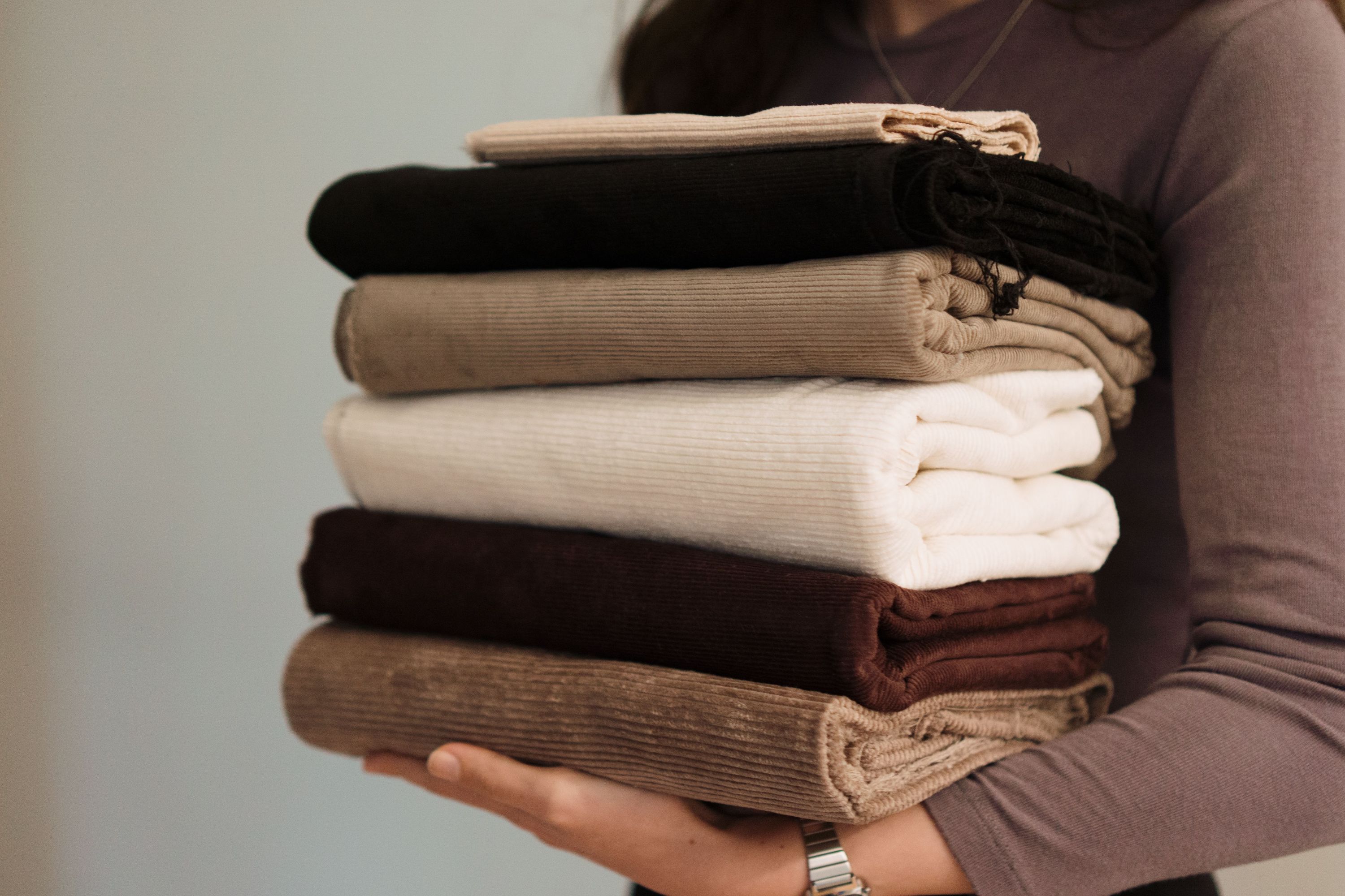 A person holding a stack of folded corduroy fabrics featuring neutral earth tones including beige, brown, and white.