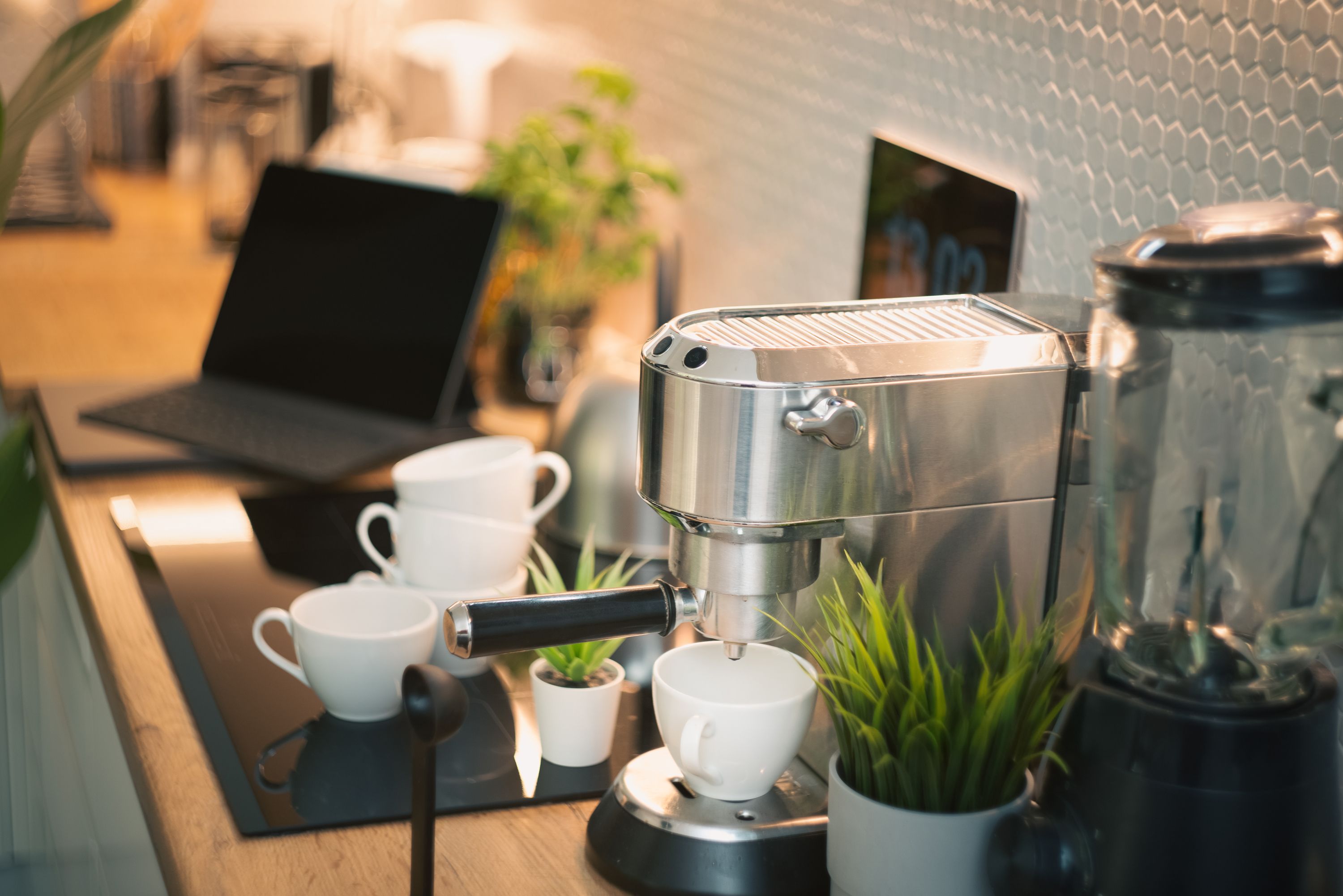 A stainless steel espresso machine resting over a small white cup on a counter next to a laptop and small potted plants