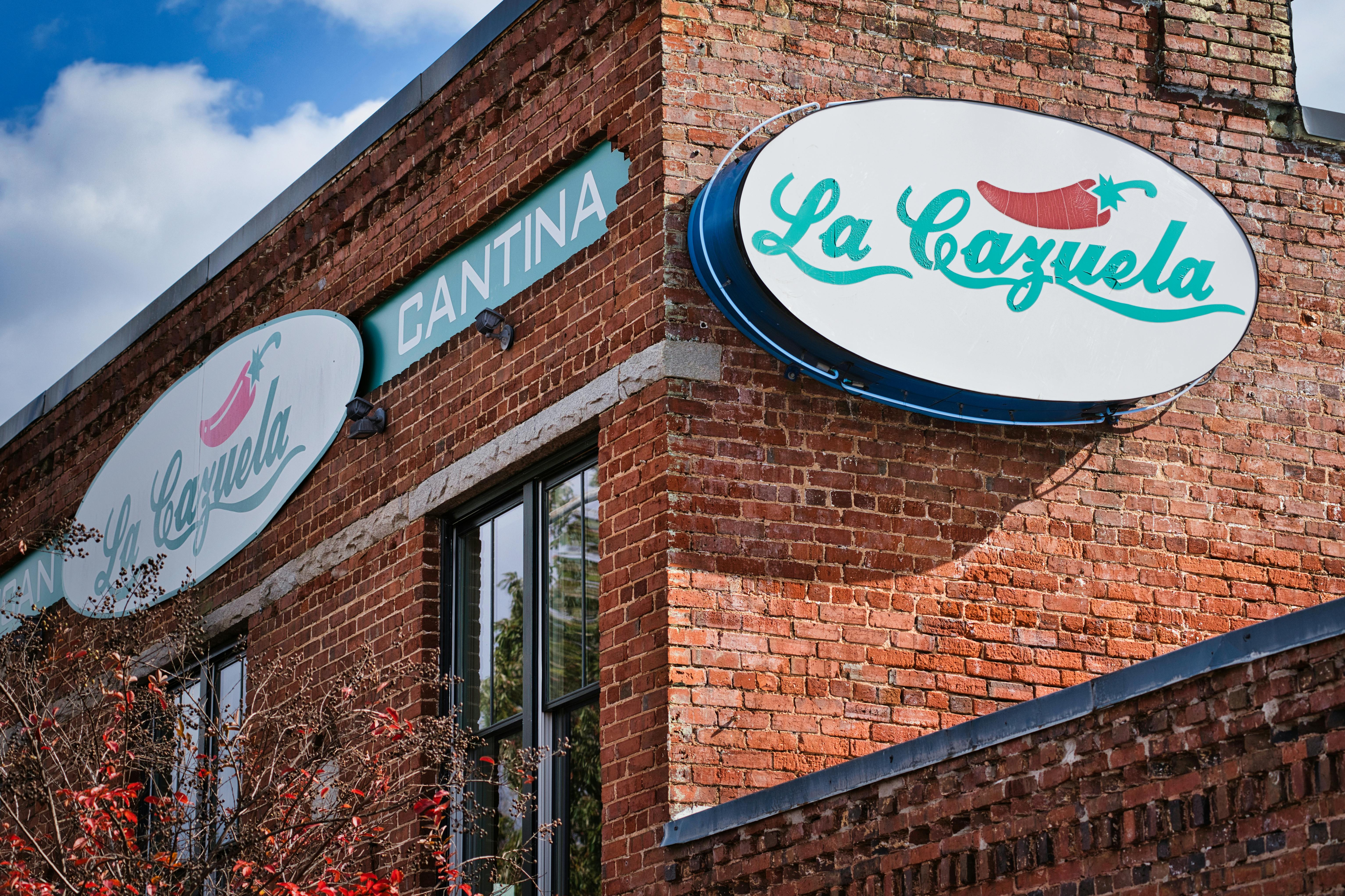 Two oval signs reading La Cazuela Cantina displaying red peppers hanging on a brick building under a blue sky.
