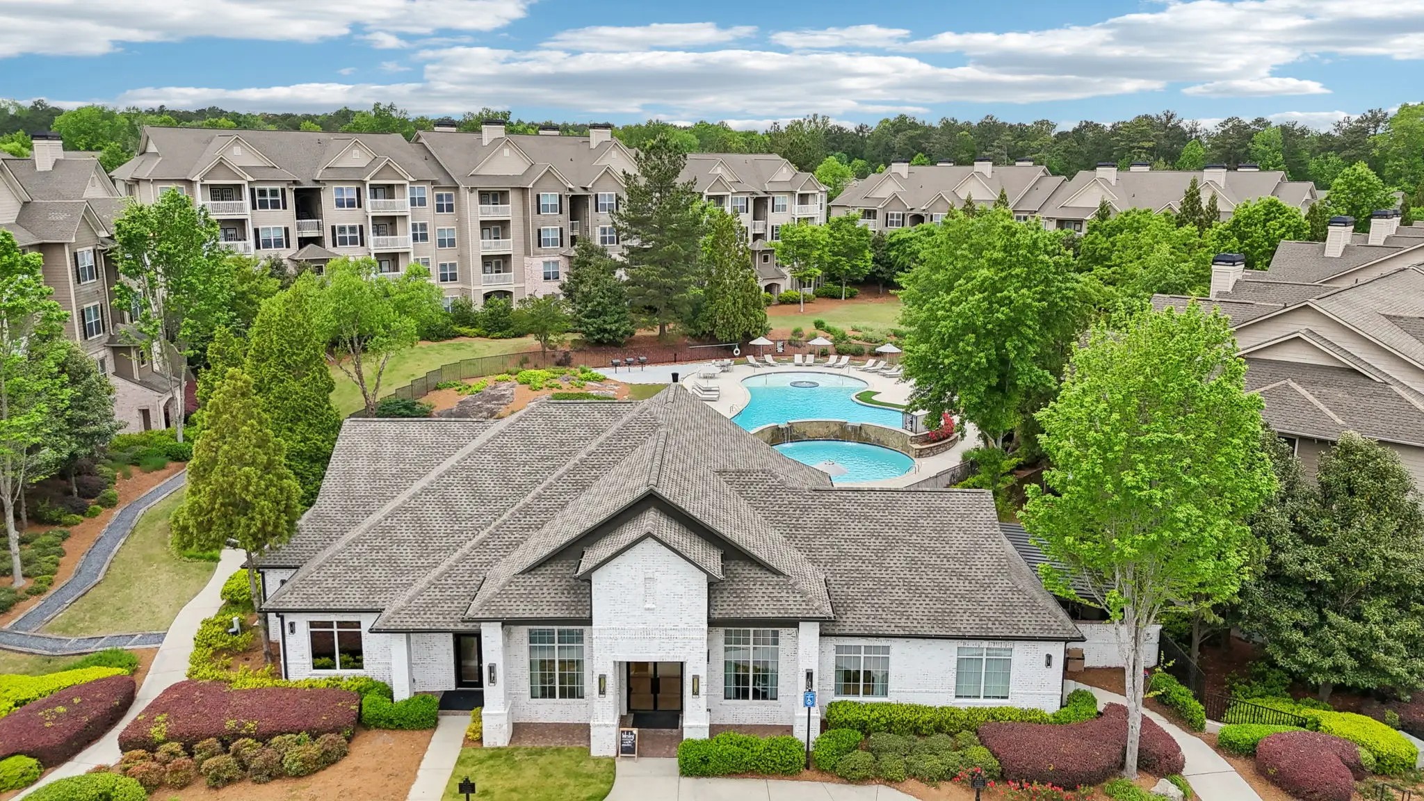 An apartment leasing office featuring a dark roof sitting near a blue swimming pool and apartment buildings among trees.