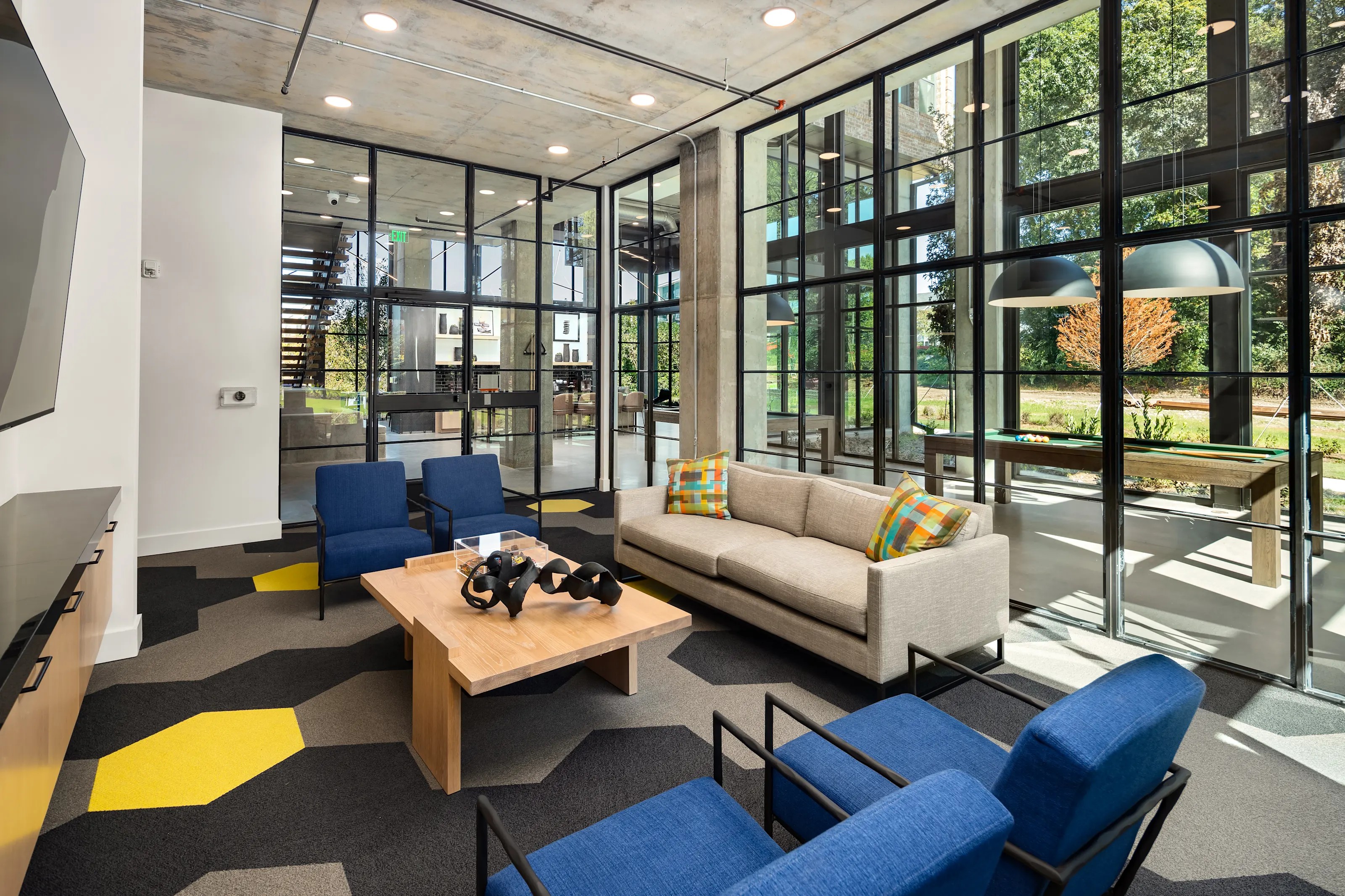 Blue armchairs and a light sofa sitting on a patterned rug near large glass windows inside The Argos Apartments lounge.