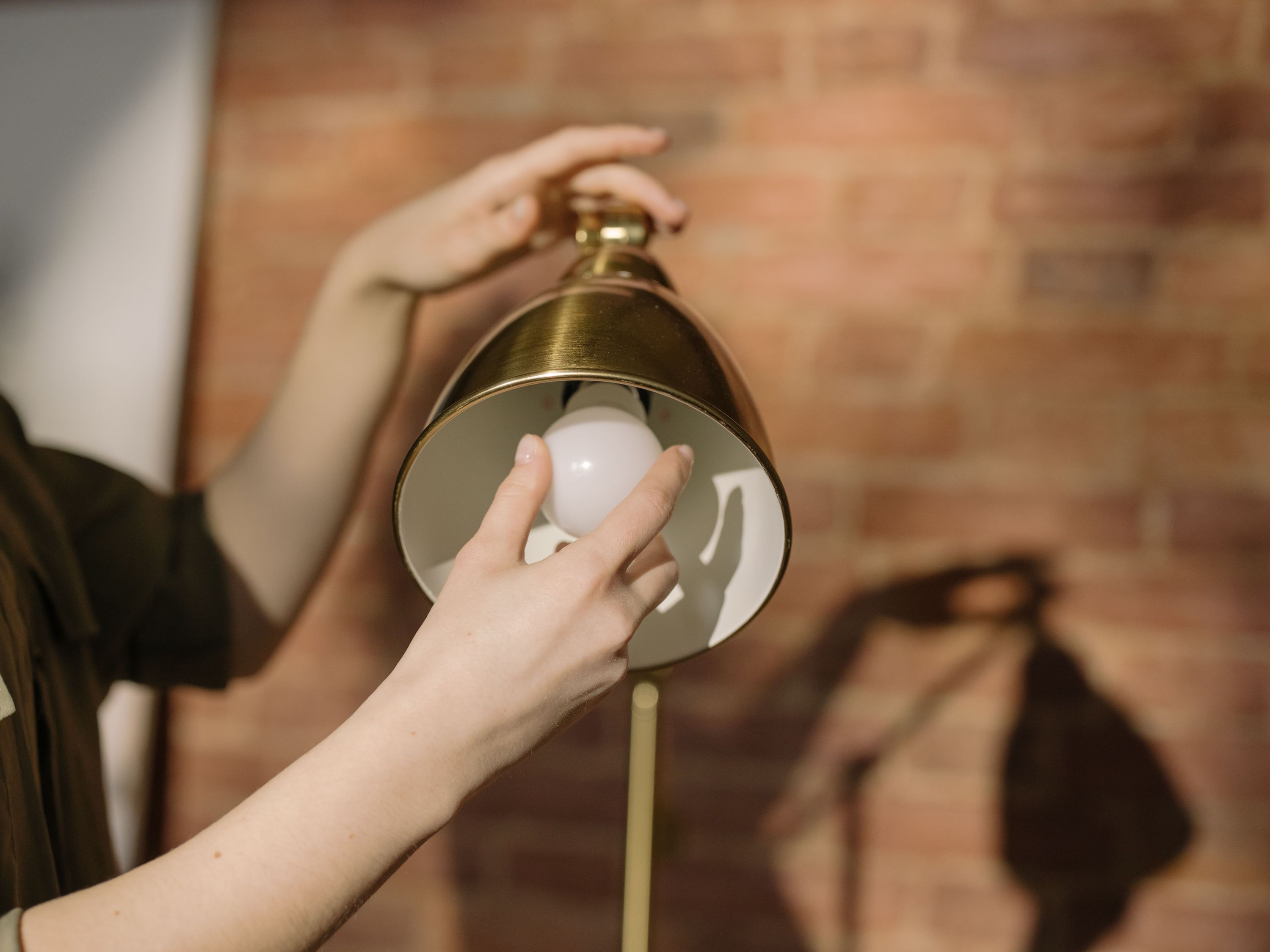 A person screwing a light bulb into a vintage brass desk lamp shade against a warm exposed brick wall background.