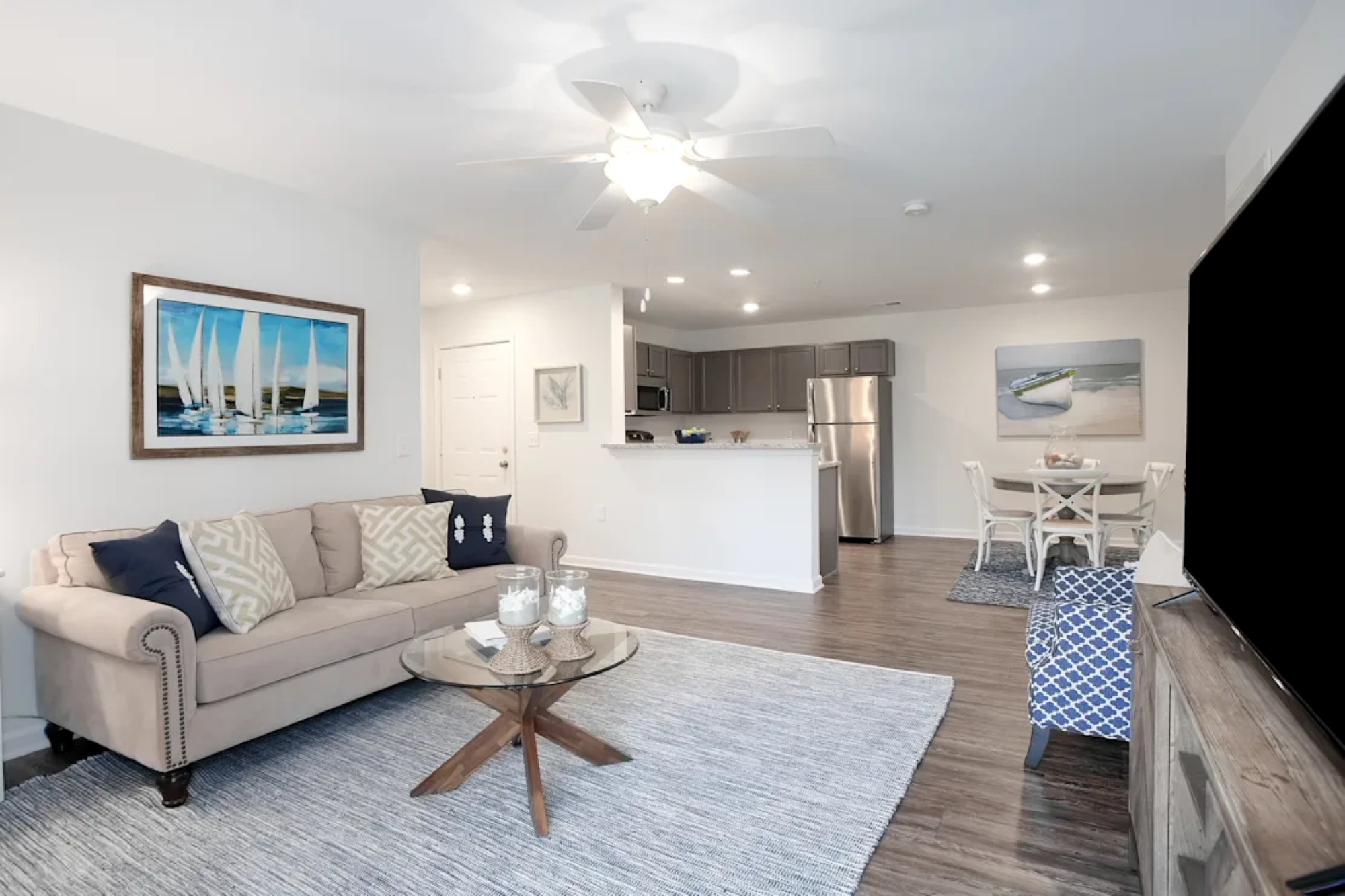 An open living room featuring a beige sofa, glass table, blue sailboat artwork, and a kitchen with dining table behind.