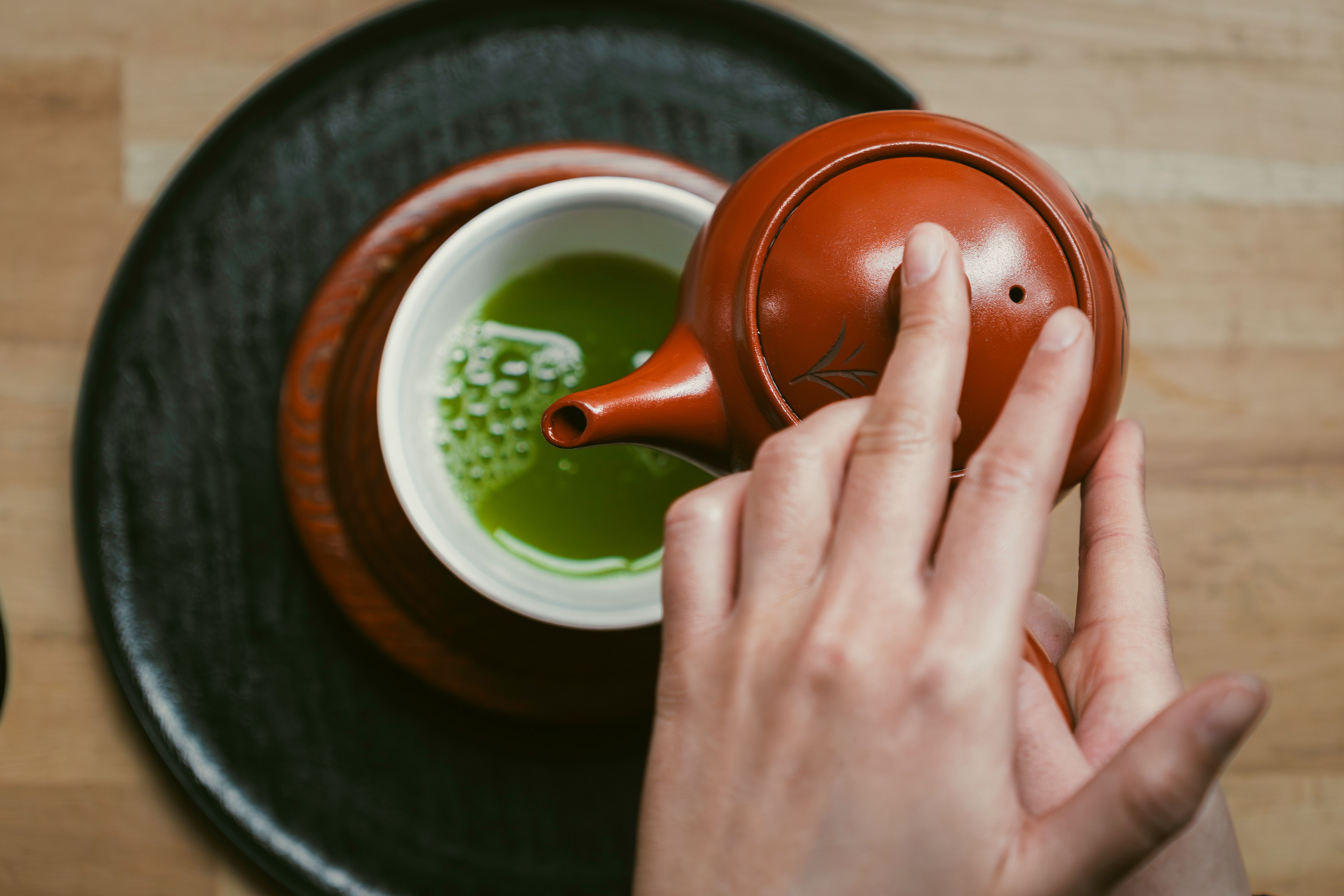 A top-down view showing a hand pouring fresh green tea from a clay teapot into a white ceramic cup on a tray.