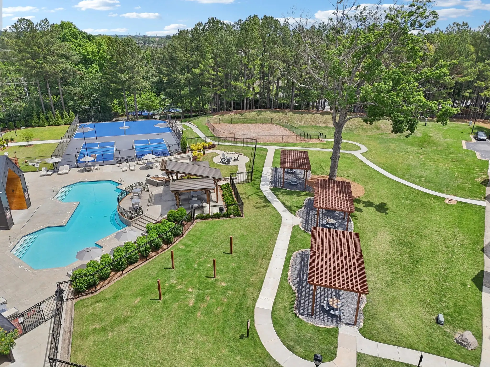 A bright blue swimming pool sitting next to blue tennis courts and wooden pavilions surrounded by trees and green grass.