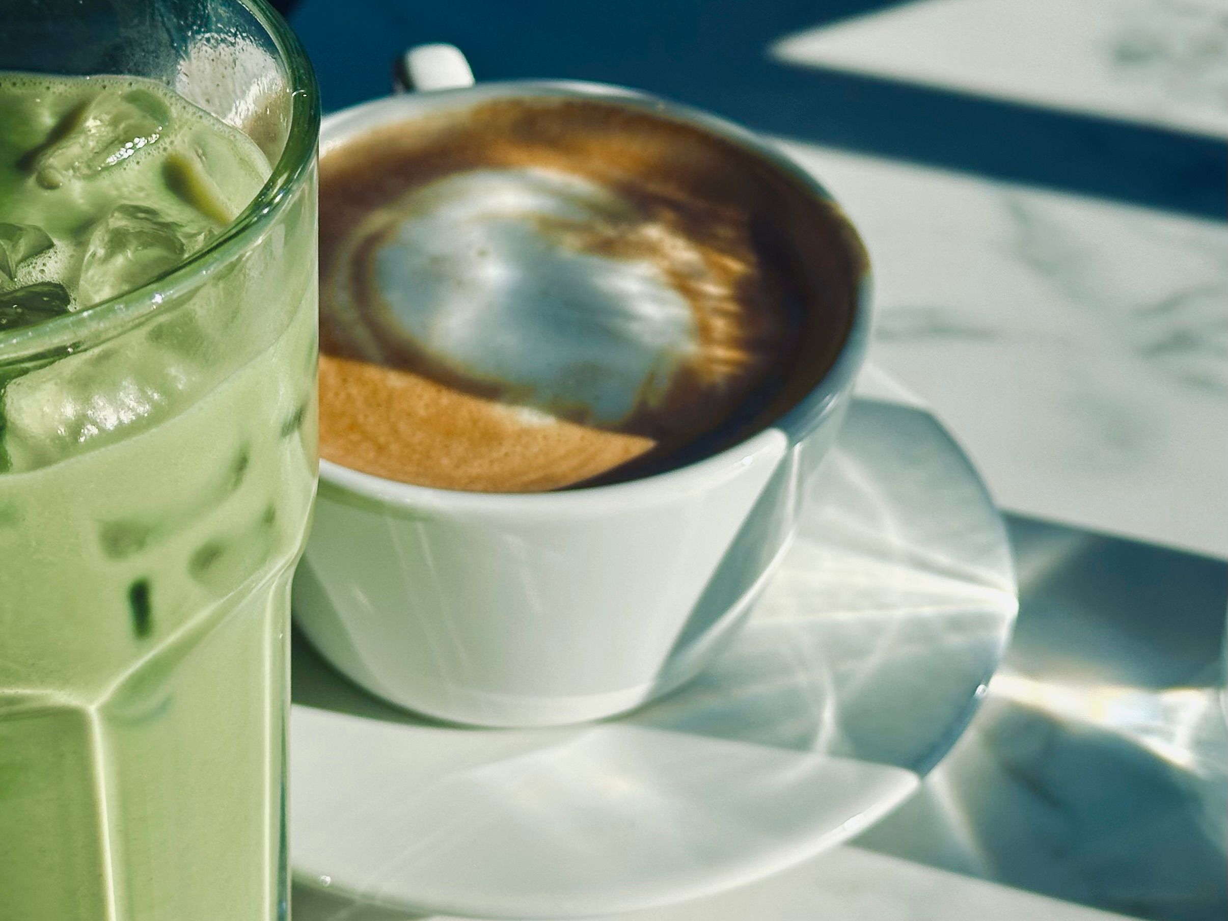 An iced matcha drink in a tall glass sitting next to a white cup of coffee on a saucer under bright sunlight on a table