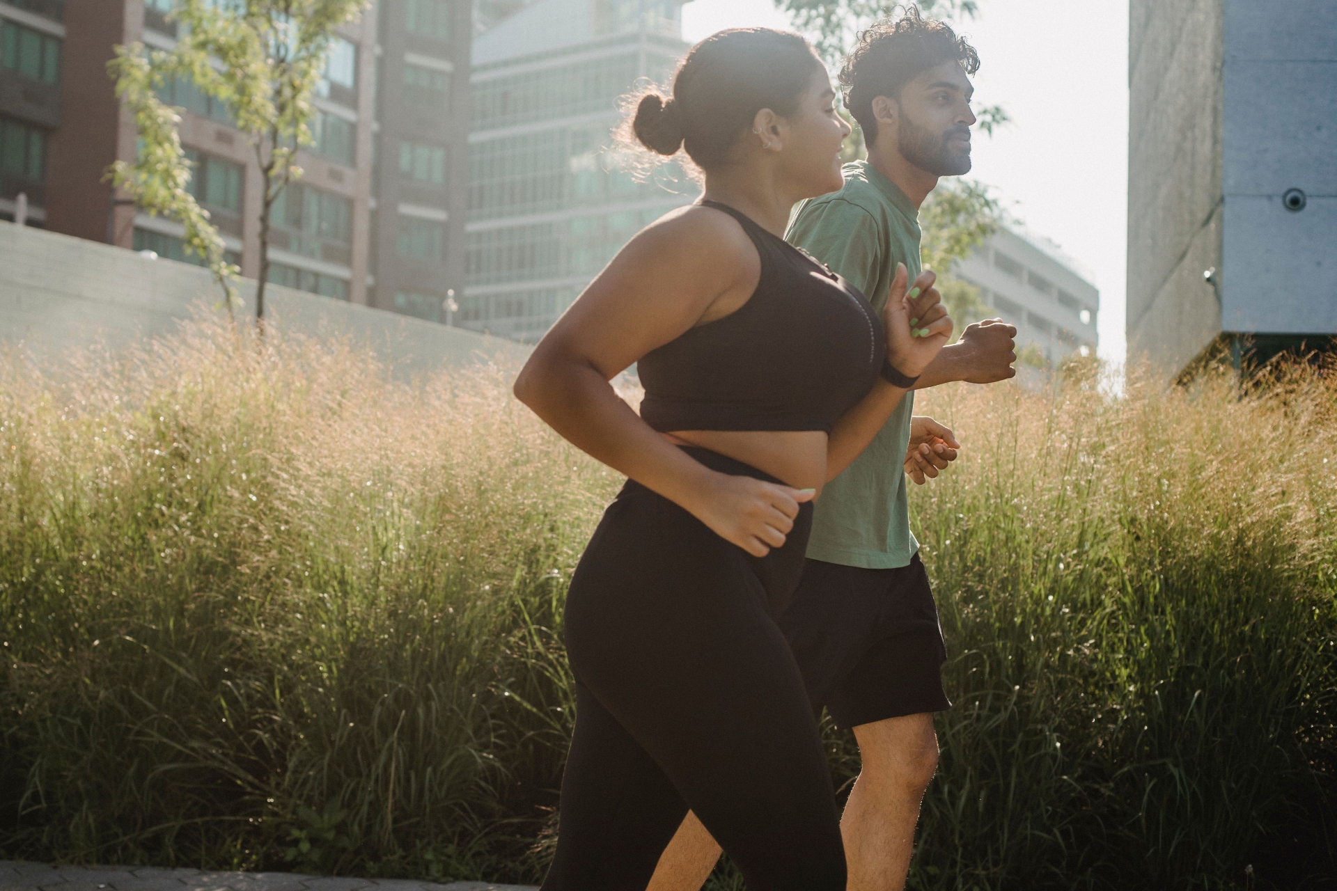 A woman wearing a black sports bra and leggings running alongside a man wearing a green shirt near tall grass.