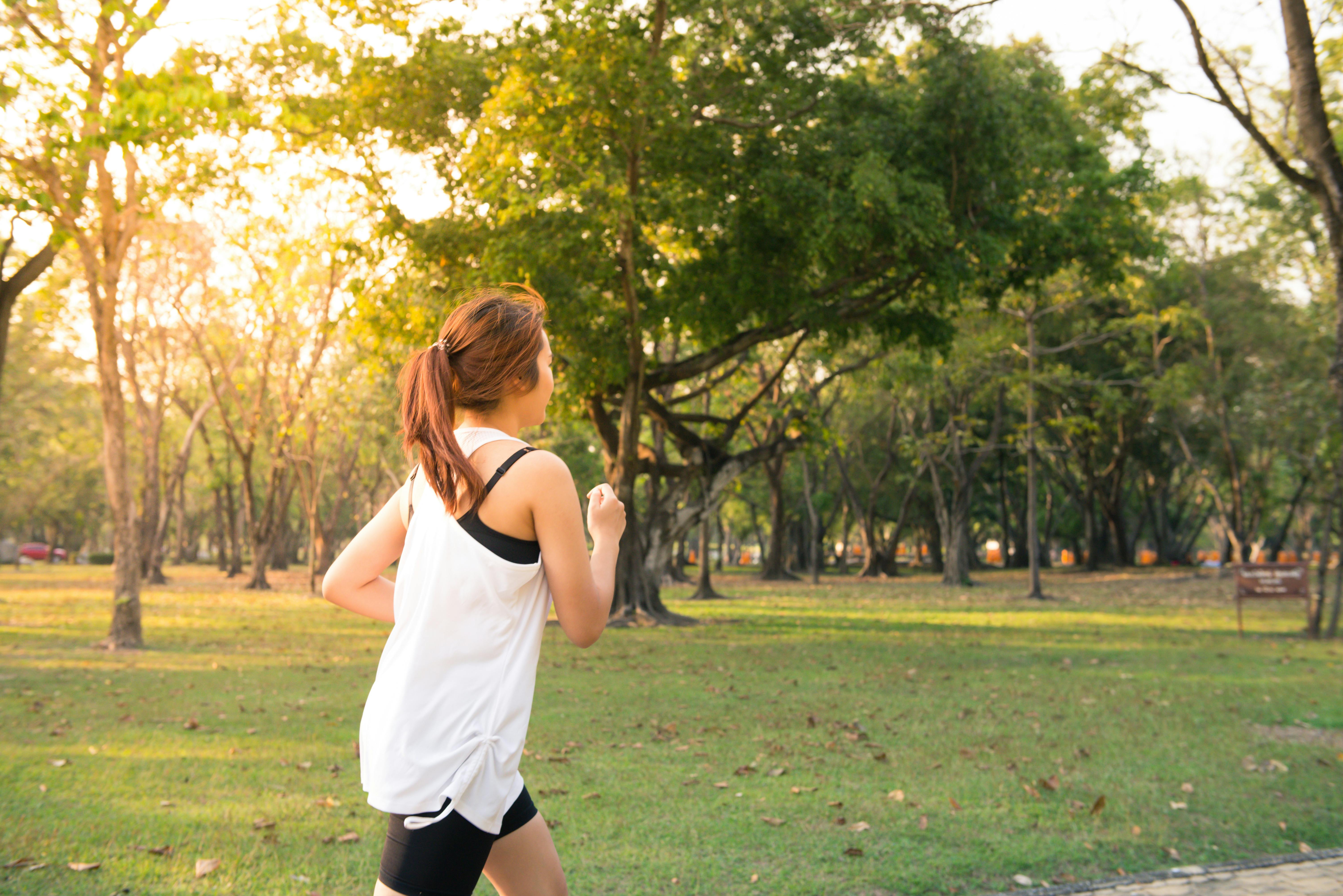 A person jogging through a sunlit park with tall trees, wearing athletic clothes and moving across grassy ground.