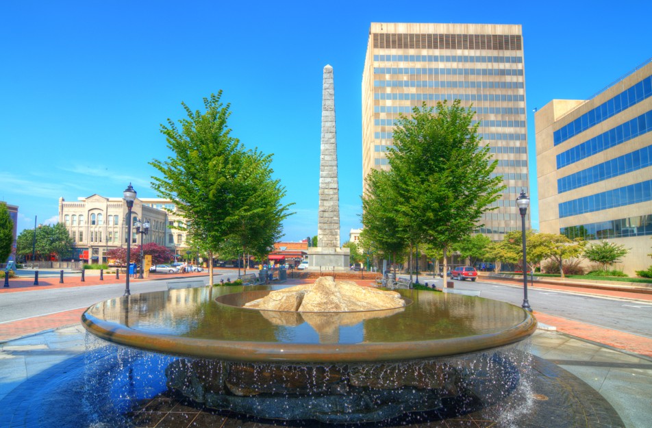 Pack Square in downtown Asheville, NC, features a fountain, trees, and an obelisk monument surrounded by buildings.