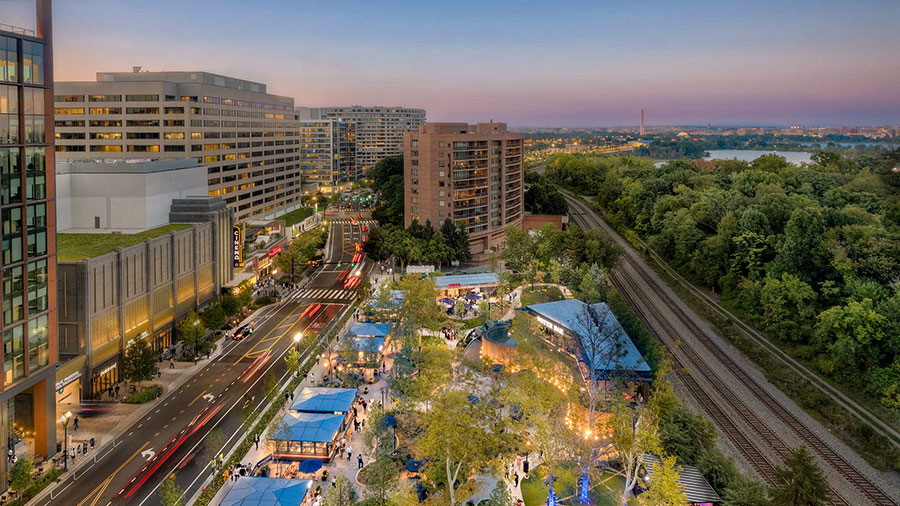 Aerial dusk view of National Landing in Arlington, VA, showing the Crystal Drive streetscape, outdoor gathering spaces, Metro rail corridor, and Reagan National Airport in the distance